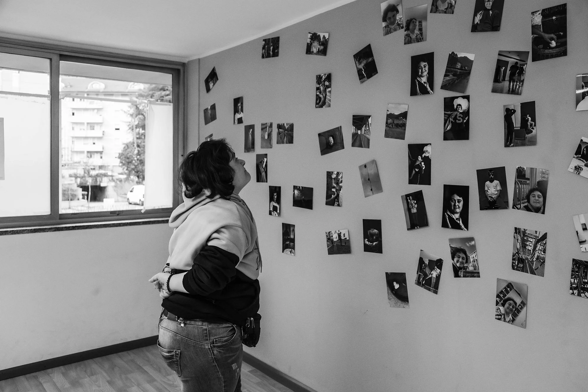 Una donna guarda un muro decorato con numerose fotografie di varie dimensioni, illuminata dalla luce proveniente da una finestra vicina.