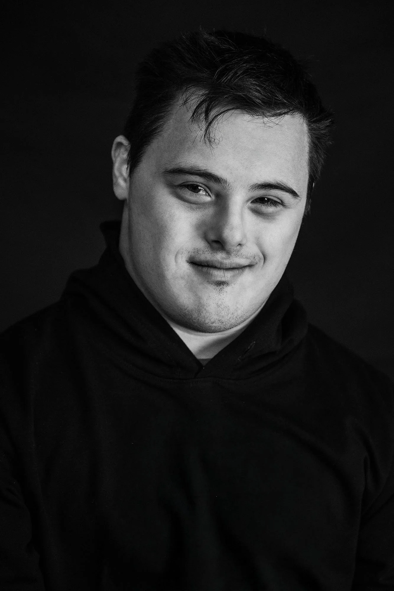Ritratto di un giovane uomo con capelli scuri e sorriso leggero, vestito con una felpa nera, sfondo nero.