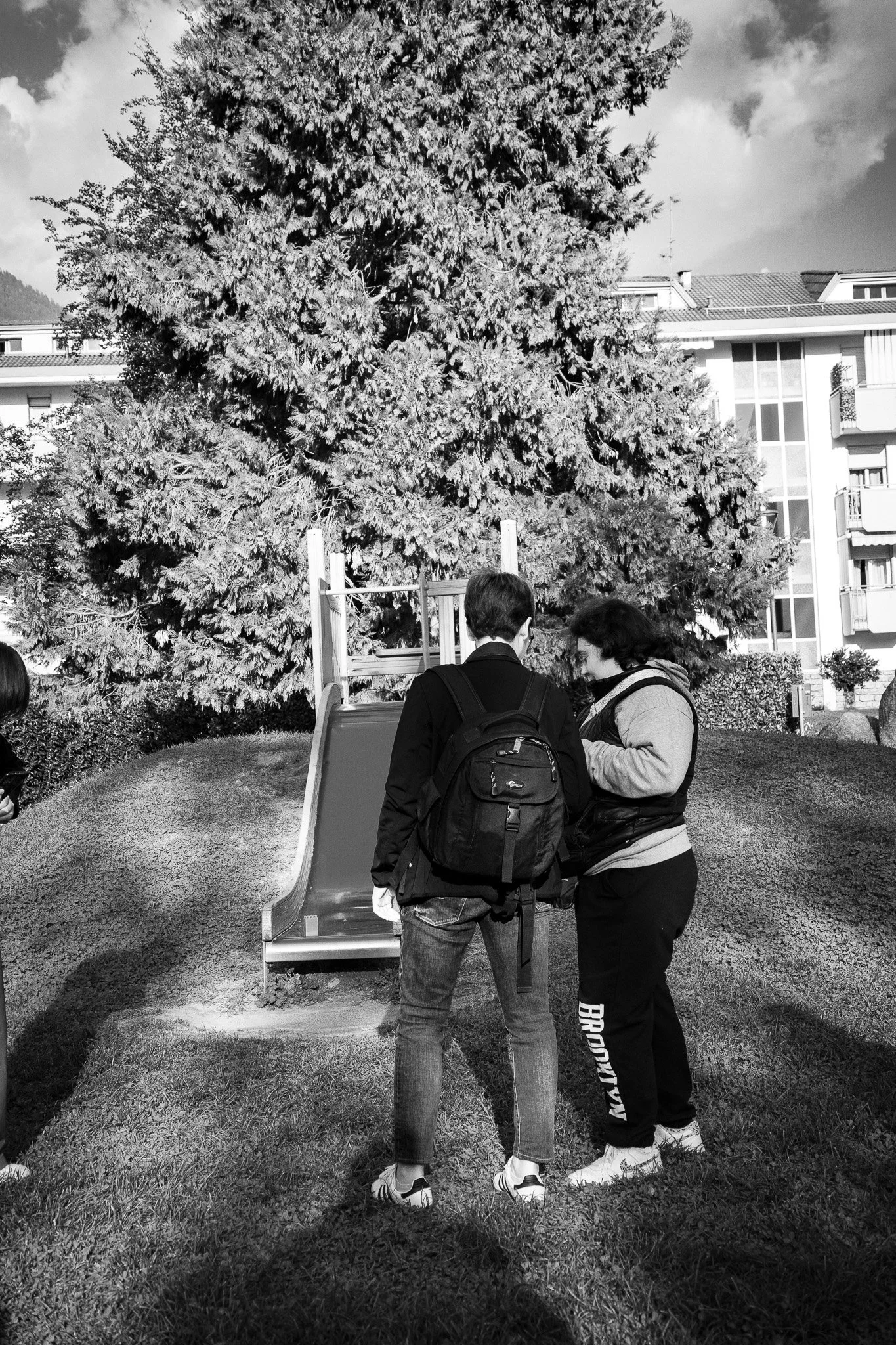 Due persone si trovano vicino a uno scivolo in un parco, con un grande albero e edifici moderni sullo sfondo.