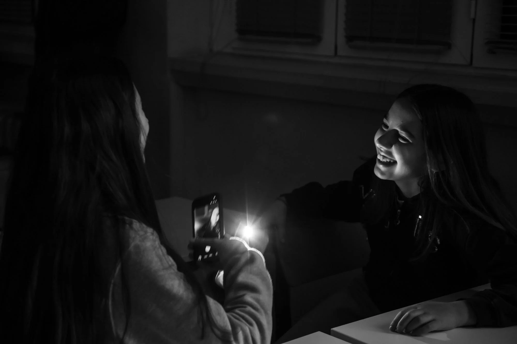 Due ragazze sorridenti sedute a un tavolo, una con un telefono che cattura il momento e l'altra con il viso illuminato da una luce, in un ambiente scolastico.