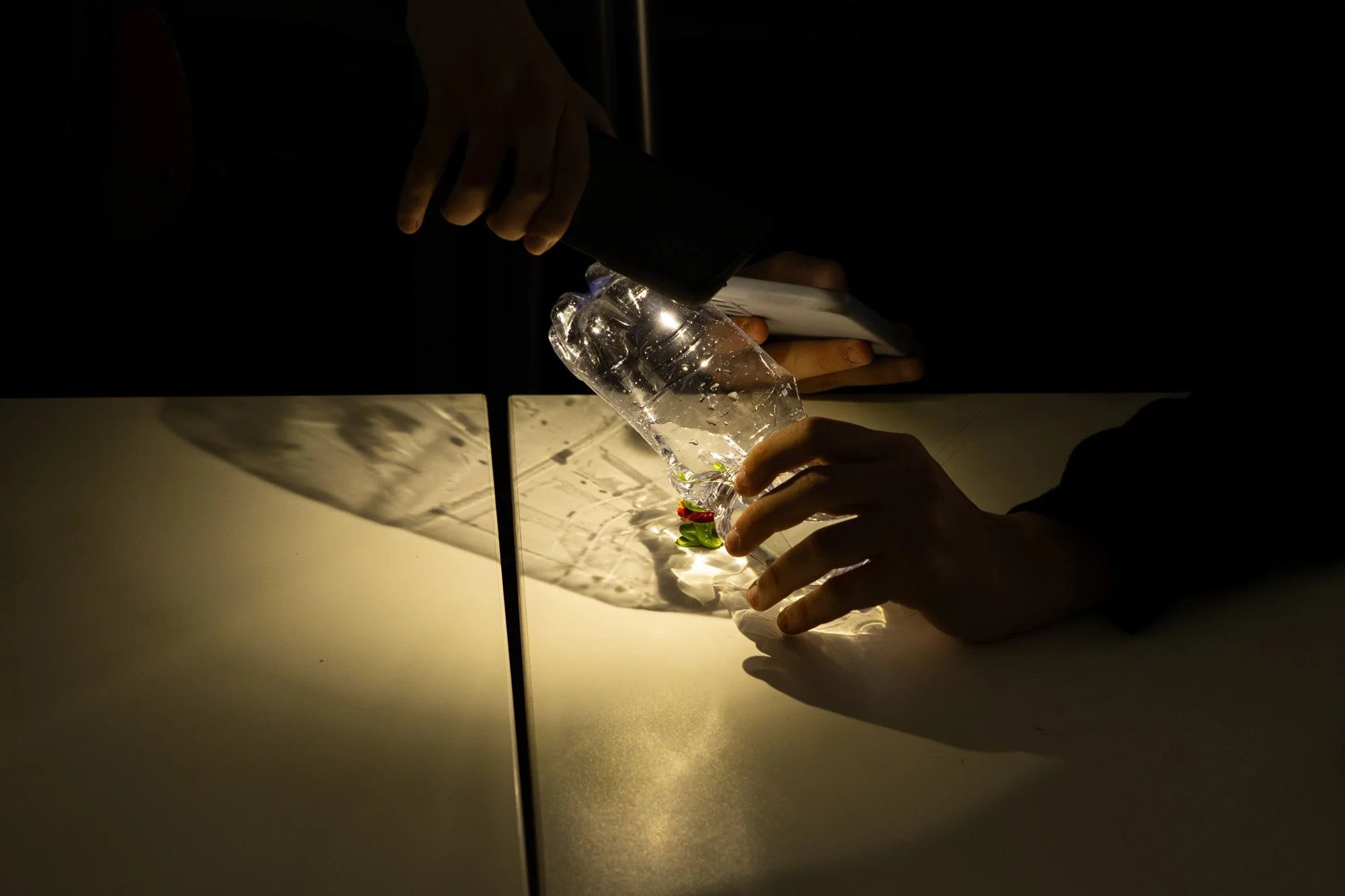 Persone che creano effetti di luce con una bottiglia di plastica, durante una lezione in classe, sulla luce in fotografia. La scena è illuminata da una luce calda con ombre su una superficie bianca.