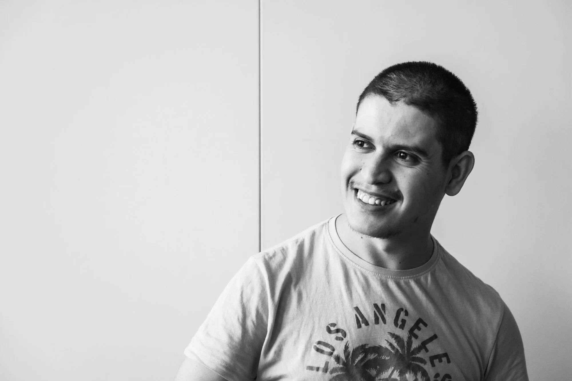Ragazzo sorridente con capelli corti, indossa una maglietta con scritte e un disegno di palme, davanti a una parete bianca.