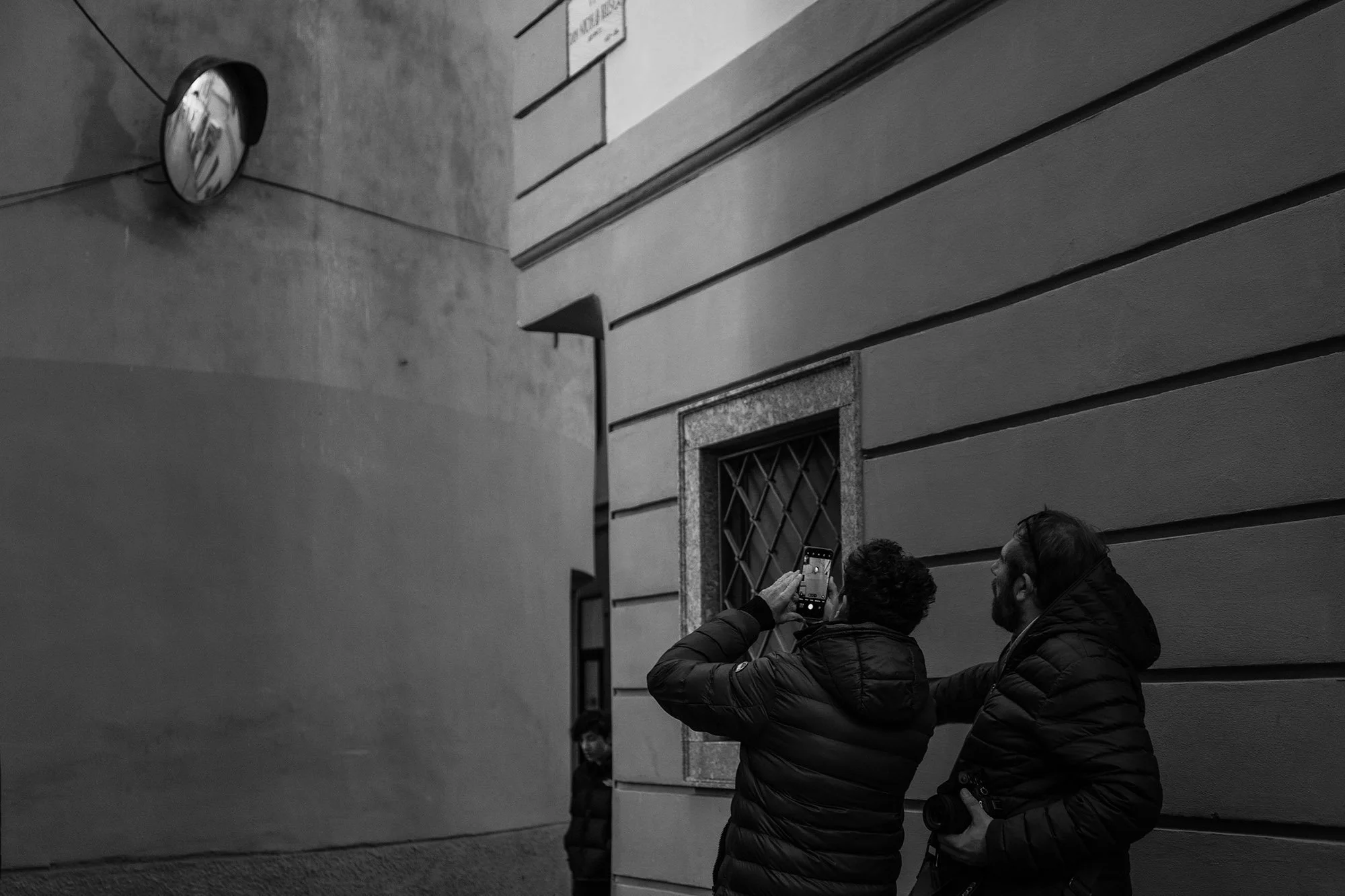 Due uomini che indossano giacche imbottite stanno scattando una foto con uno smartphone, vicino a una parete di un edificio, in una scena all'aperto.