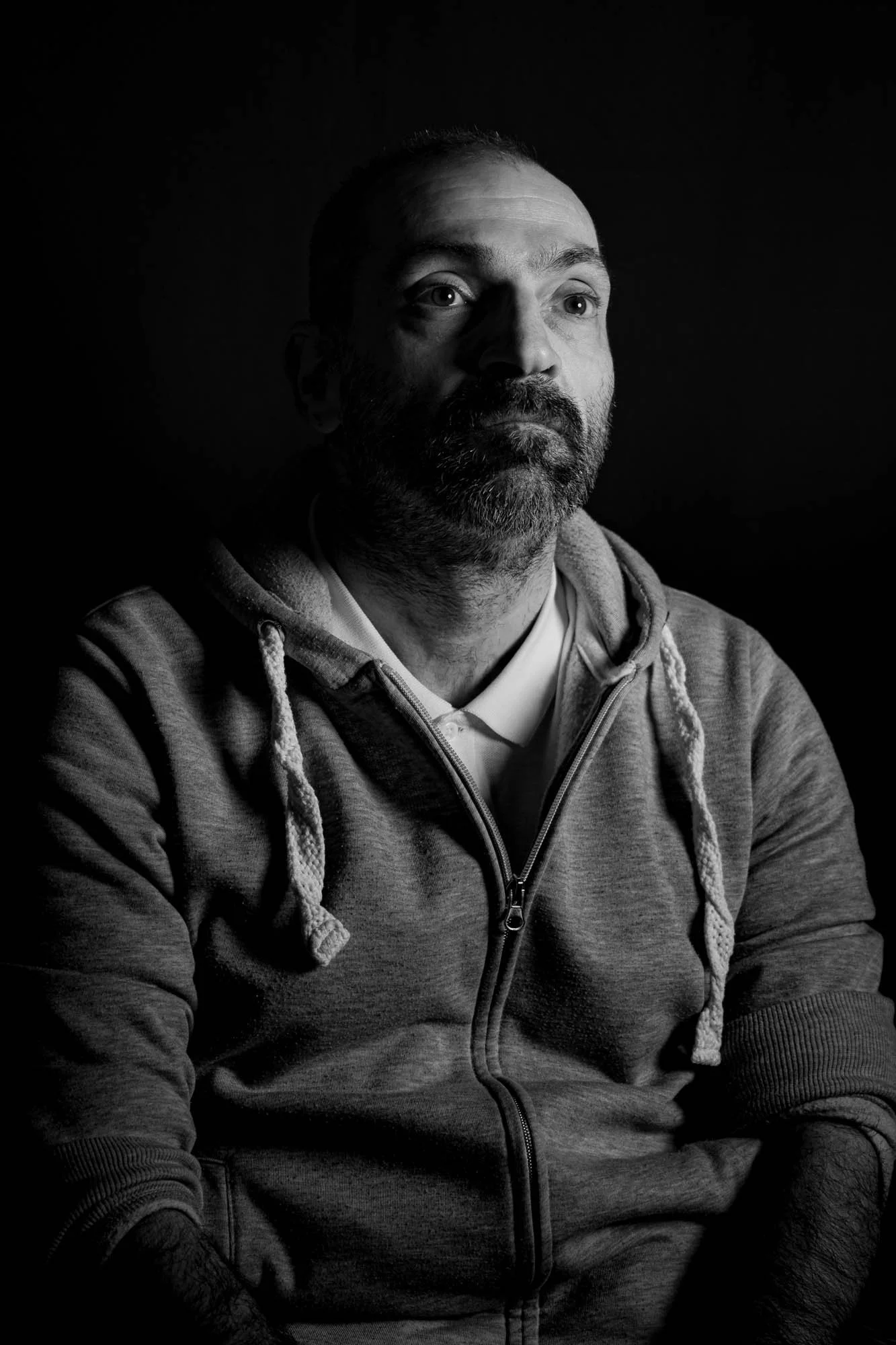 Un uomo con barba indossa una felpa grigia con cappuccio e si trova contro uno sfondo nero, in un'immagine in bianco e nero.