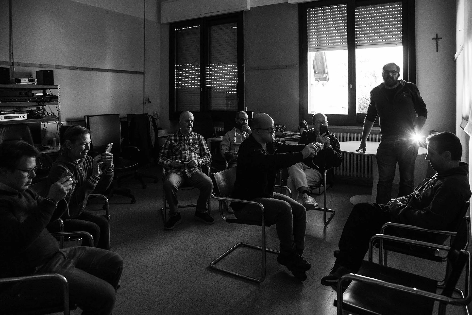 Un gruppo di persone sedute in una stanza che partecipano ad una lezione di fotografia, alcune che usano smartphone, con un uomo che si trova in piedi e illumina con la torcia un uomo seduto. Da una finestra aperta alle sue spalle entra la luce natur