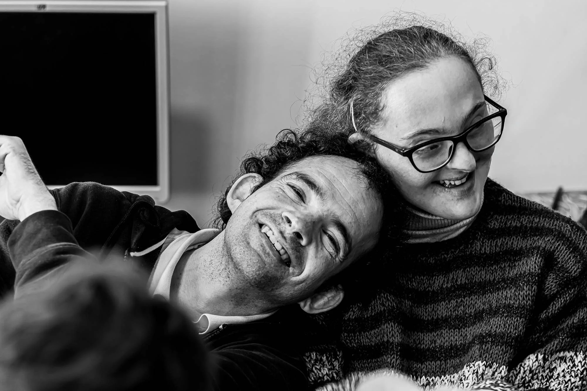 Due persone sorridenti abbracciate, una donna con occhiali e capelli ricci, un uomo con capelli ricci e barba, che si mettono in posa per la fotocamera in un ambiente interno.