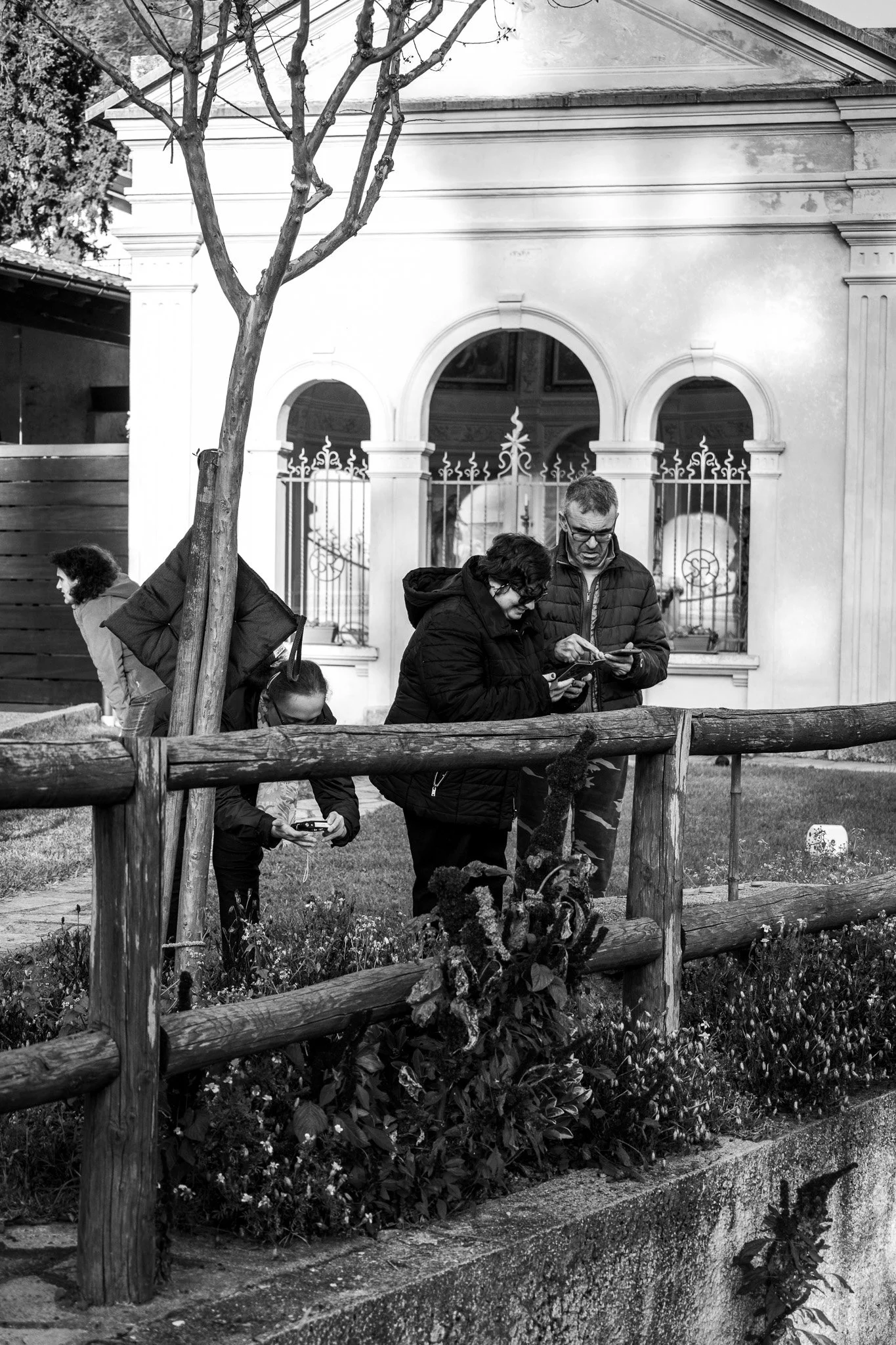 Persone che camminano in un parco, alcune utilizzano telefoni cellulari, dietro una recinzione in legno e un edificio con vetrate e archi decorativi.