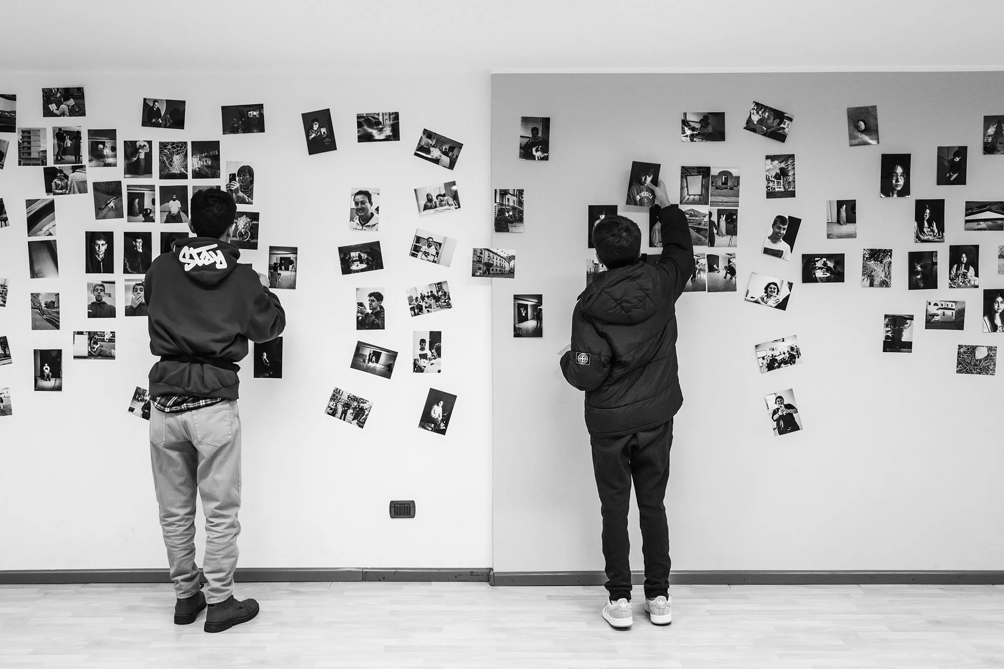 Due ragazzi, visti da dietro, osservano una parete con foto artistiche appese e appendono le loro stampe al muro. L'ambiente è un'immagine in bianco e nero.
