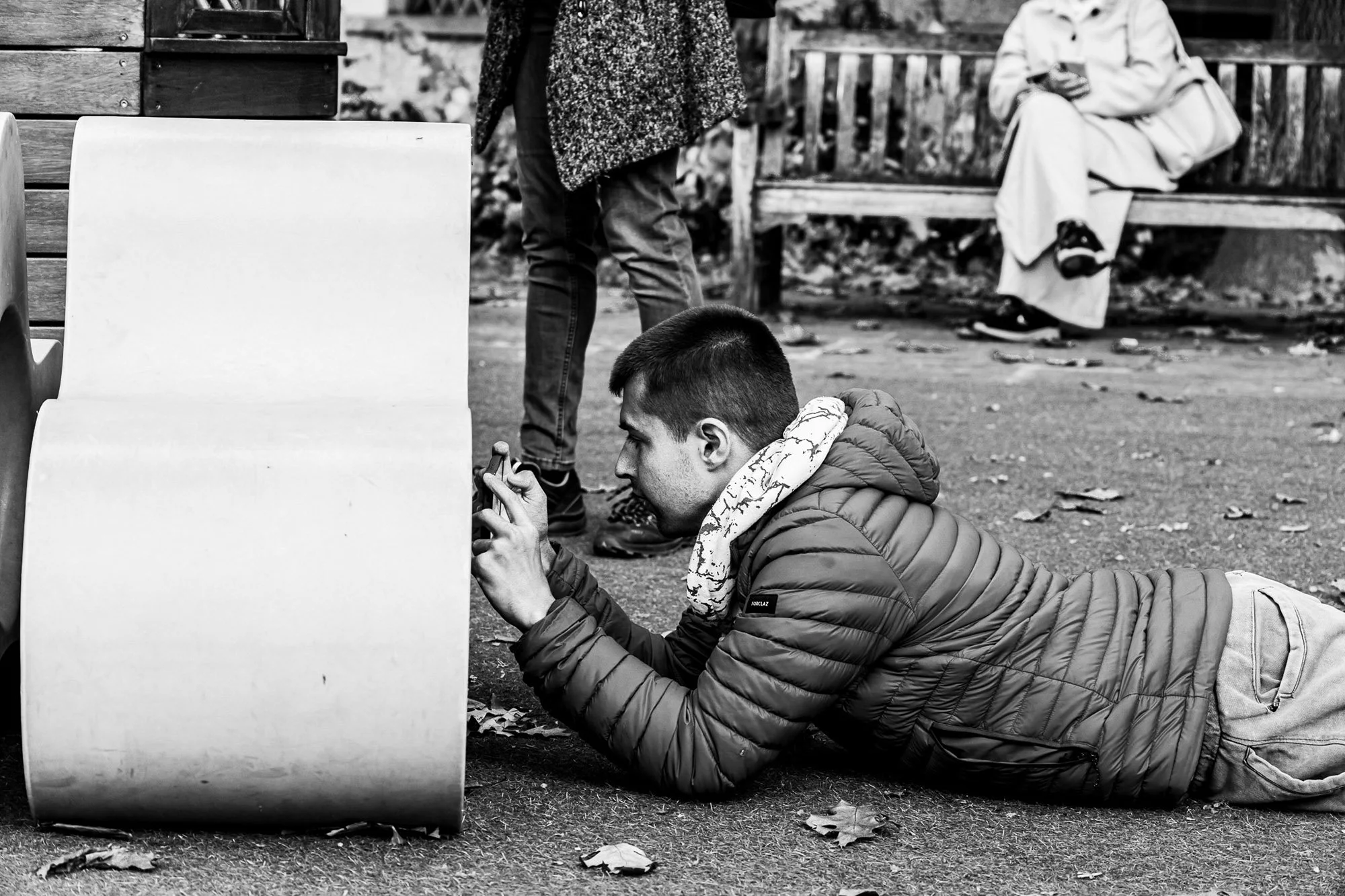 Ragazzo disteso sul suolo che utilizza uno smartphone, indossa una giacca imbottita e una sciarpa, accanto a un banco in un parco.