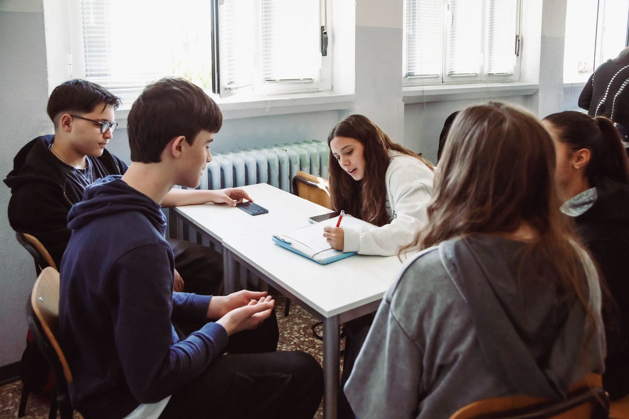 Gruppo di studenti seduti intorno a un tavolo in una classe, che discutono e prendono appunti, con finestre e luce naturale.