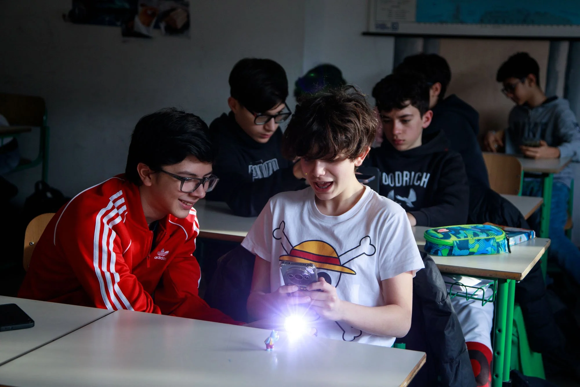 Gruppo di ragazzi in classe che guardano uno smartphone, con effetti di luce dal dispositivo.