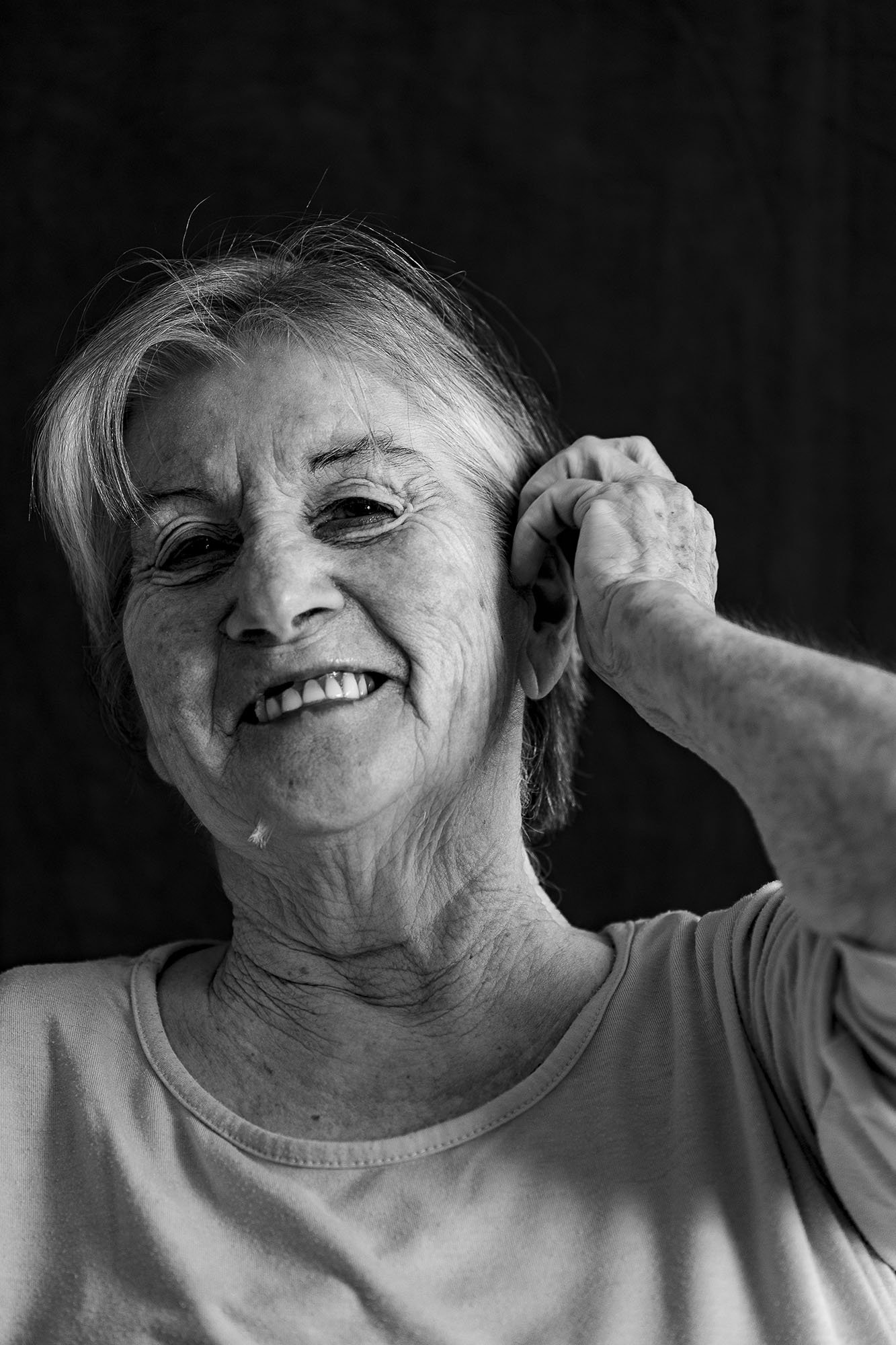 Ritratto di una donna anziana sorridente in bianco e nero, che si tocca l'orecchio con la mano destra.