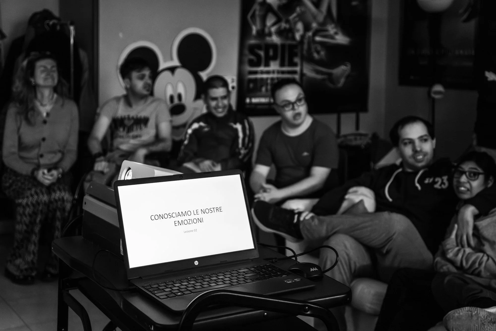 Un gruppo di persone sedute in una stanza, alcune sorridenti e altre con espressioni pensierose, sullo sfondo un poster di Topolino e una scaffalatura con oggetti vari. In primo piano, un computer portatile mostra una slide con la scritta "CONOSCIAMO