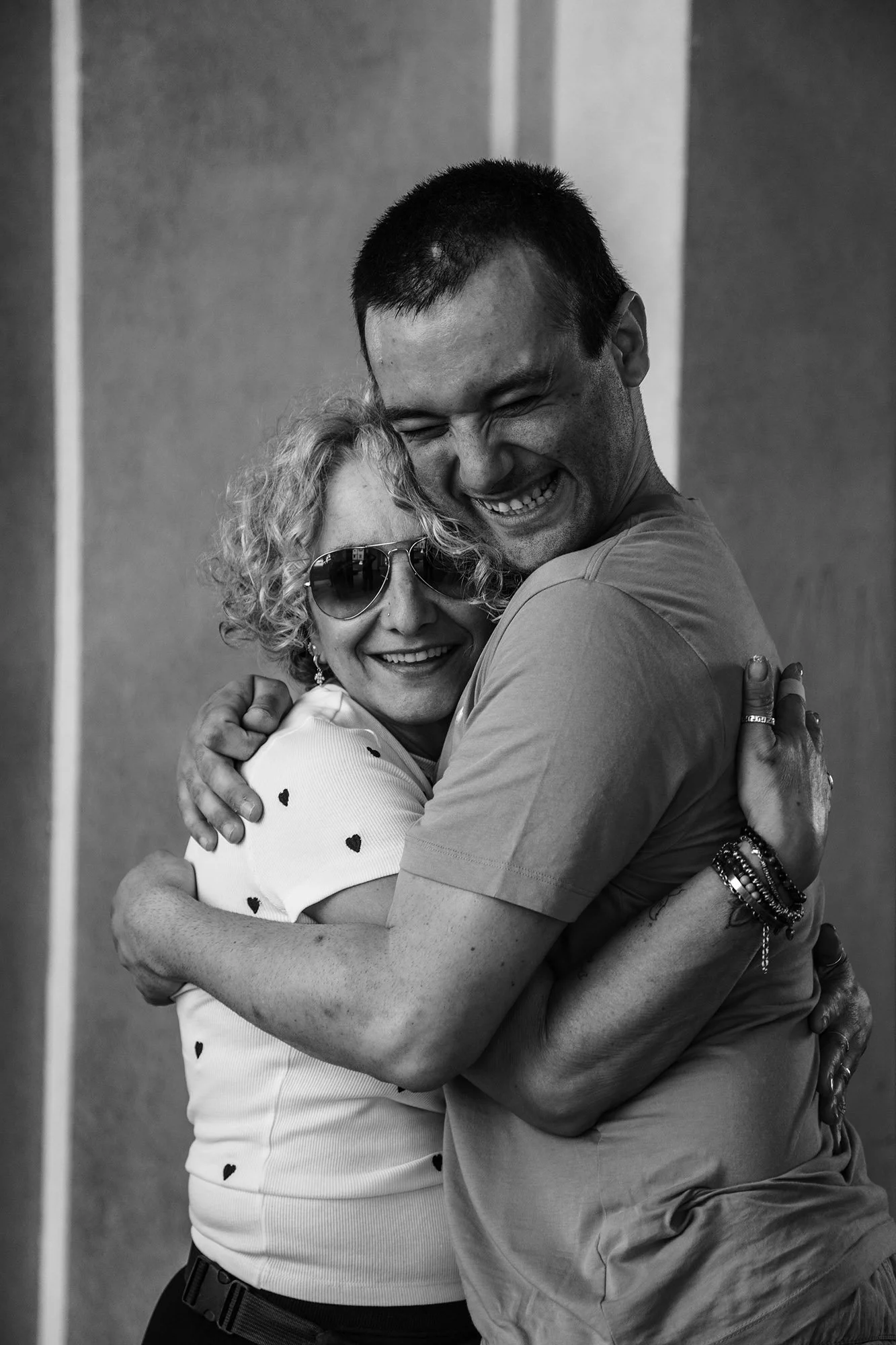 Due persone in un abbraccio stretto, sorridenti e felici, una indossa occhiali da sole e ha capelli ricci, l'altra ha i capelli corti e scuri.