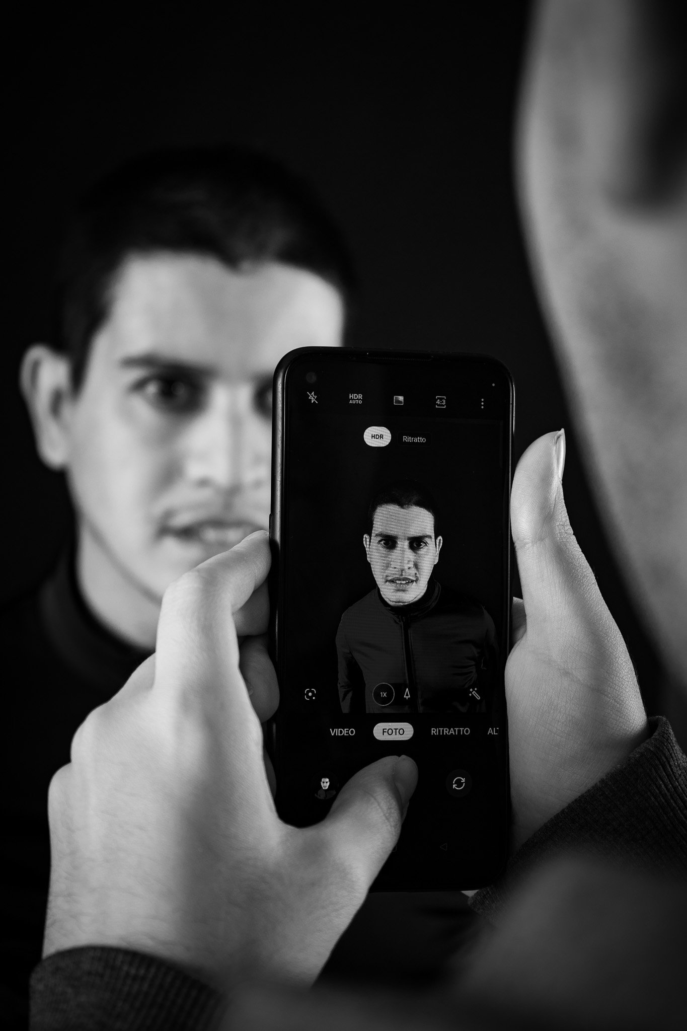 Una persona che scatta una foto con uno smartphone di un uomo con capelli scuri, sfocato sullo sfondo. La scena è in bianco e nero.