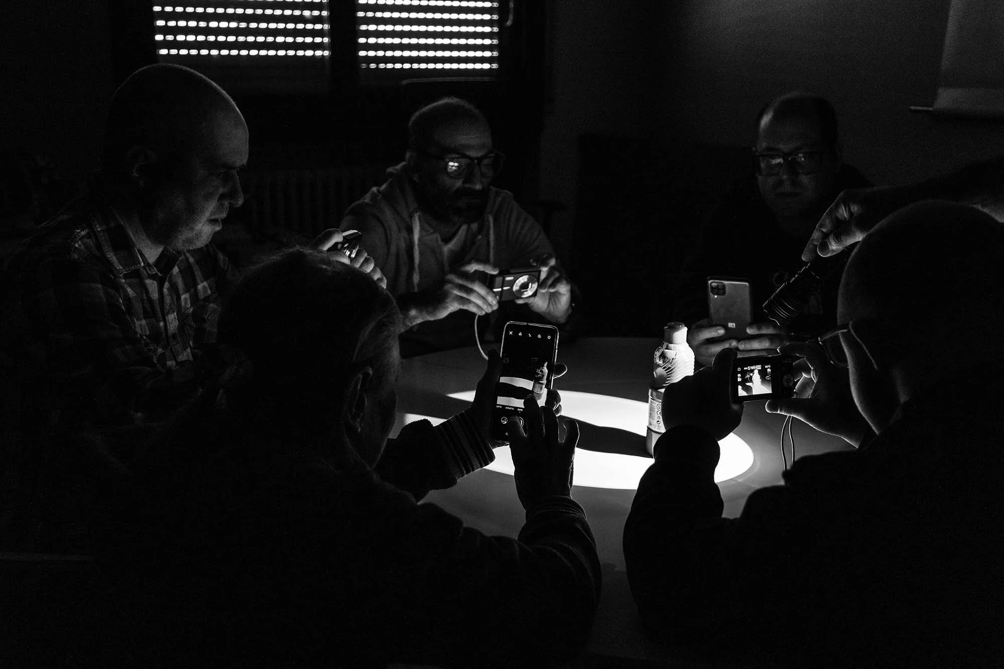 Gruppo di persone sedute intorno a un tavolo, alcune che usano smartphone e alcuni fotocamere, in un ambiente buio illuminato da una luce intensa proveniente dal centro del tavolo.