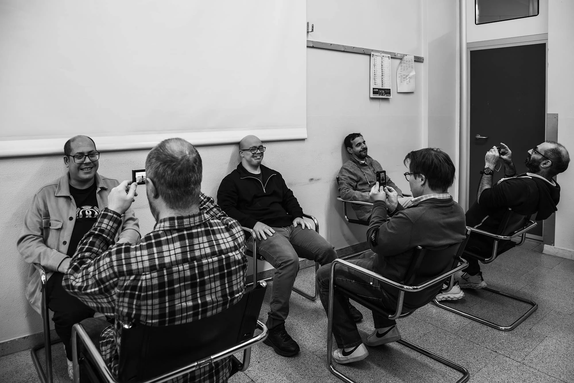 Un gruppo di persone sedute in una sala, alcune scattano foto o registrano con il cellulare mentre un uomo si sente sorridente. L'ambiente sembra una stanza di un'aula o di un ufficio.