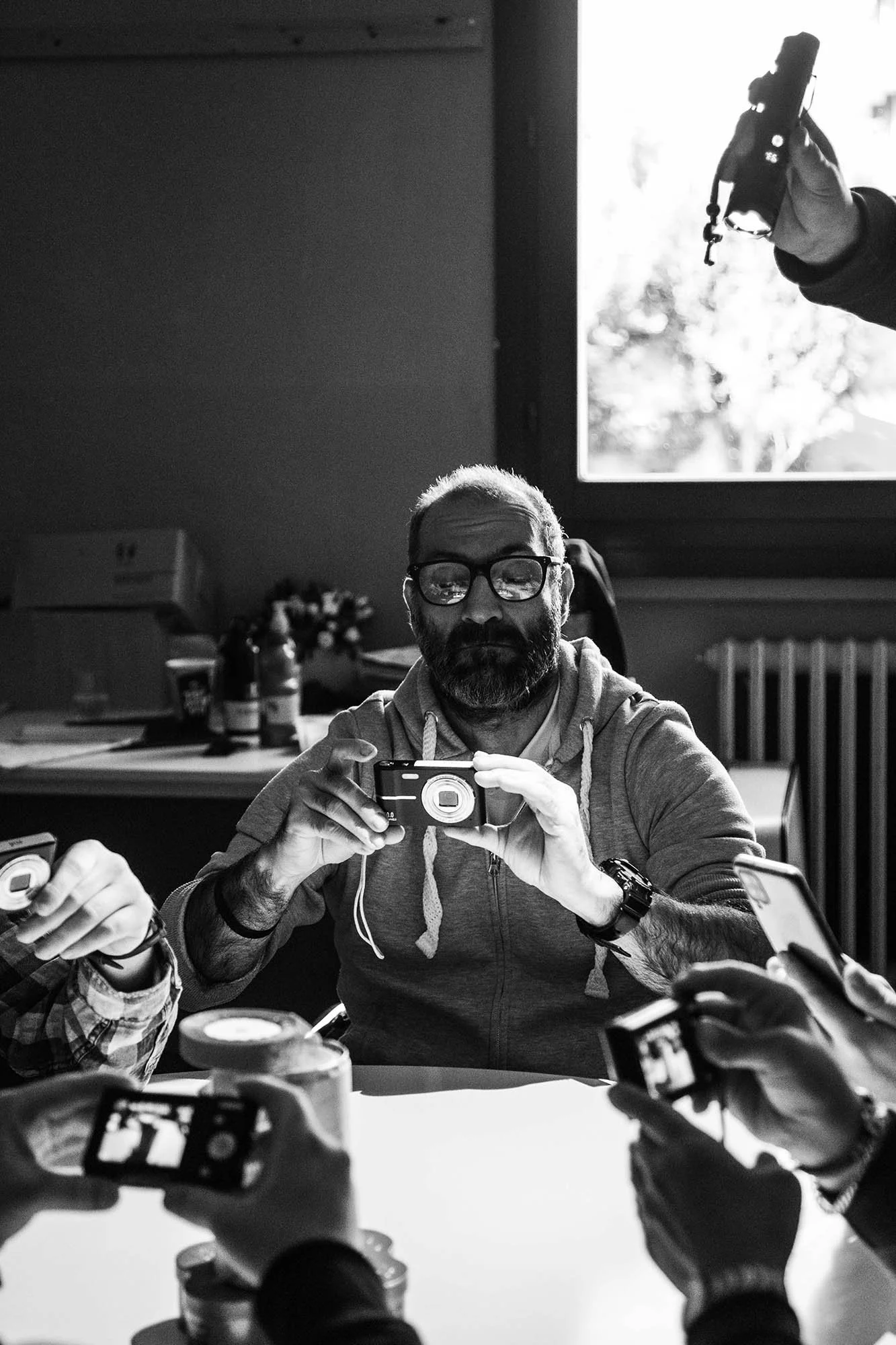 Un uomo con barba e occhiali scatta una foto di sé stesso con una fotocamera digitale, seduto a un tavolo circondato da altre persone con telecamere. Nell'ambiente, una finestra aperta mostra la luce naturale.