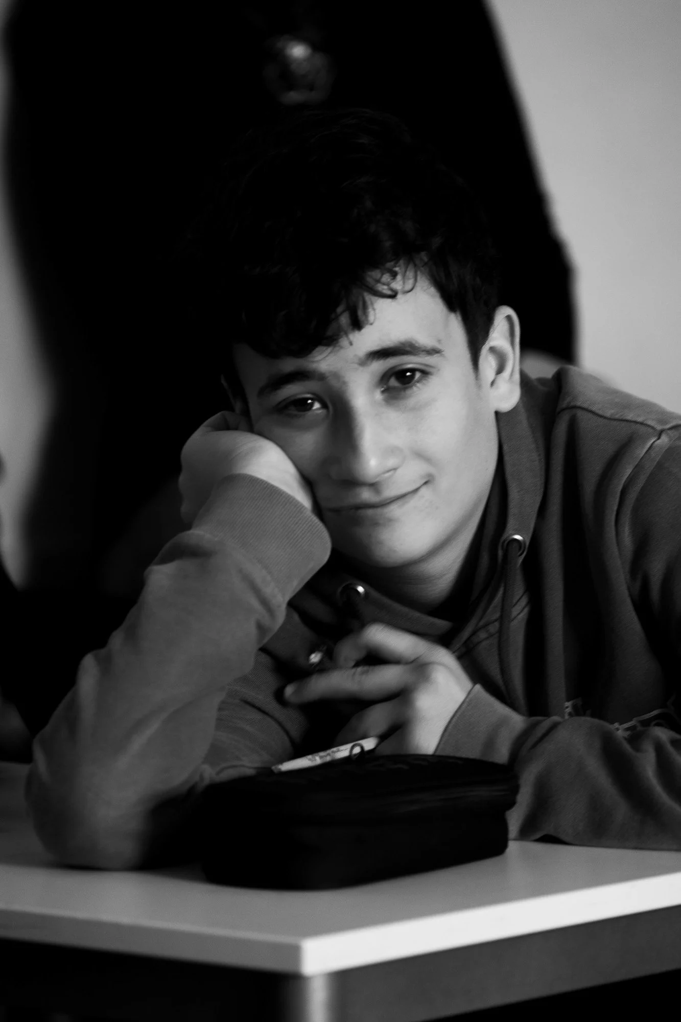 Ragazzo sorridente che guarda la fotocamera, seduto davanti a un tavolo con un cellulare in mano, con uno sfondo sfocato in bianco e nero.