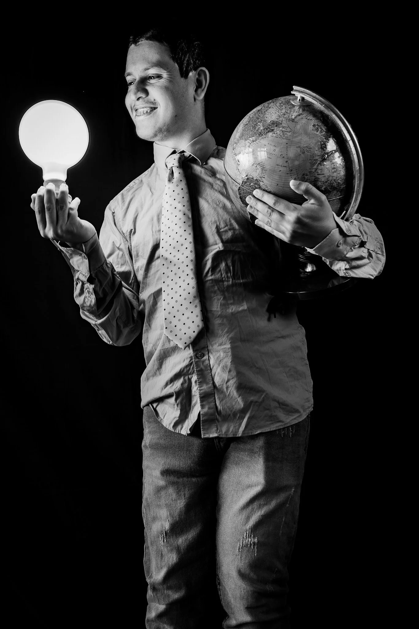 Ragazzo con camicia e cravatta che tiene in mano una lampadina accesa e una luna in miniatura, in un'immagine in bianco e nero.