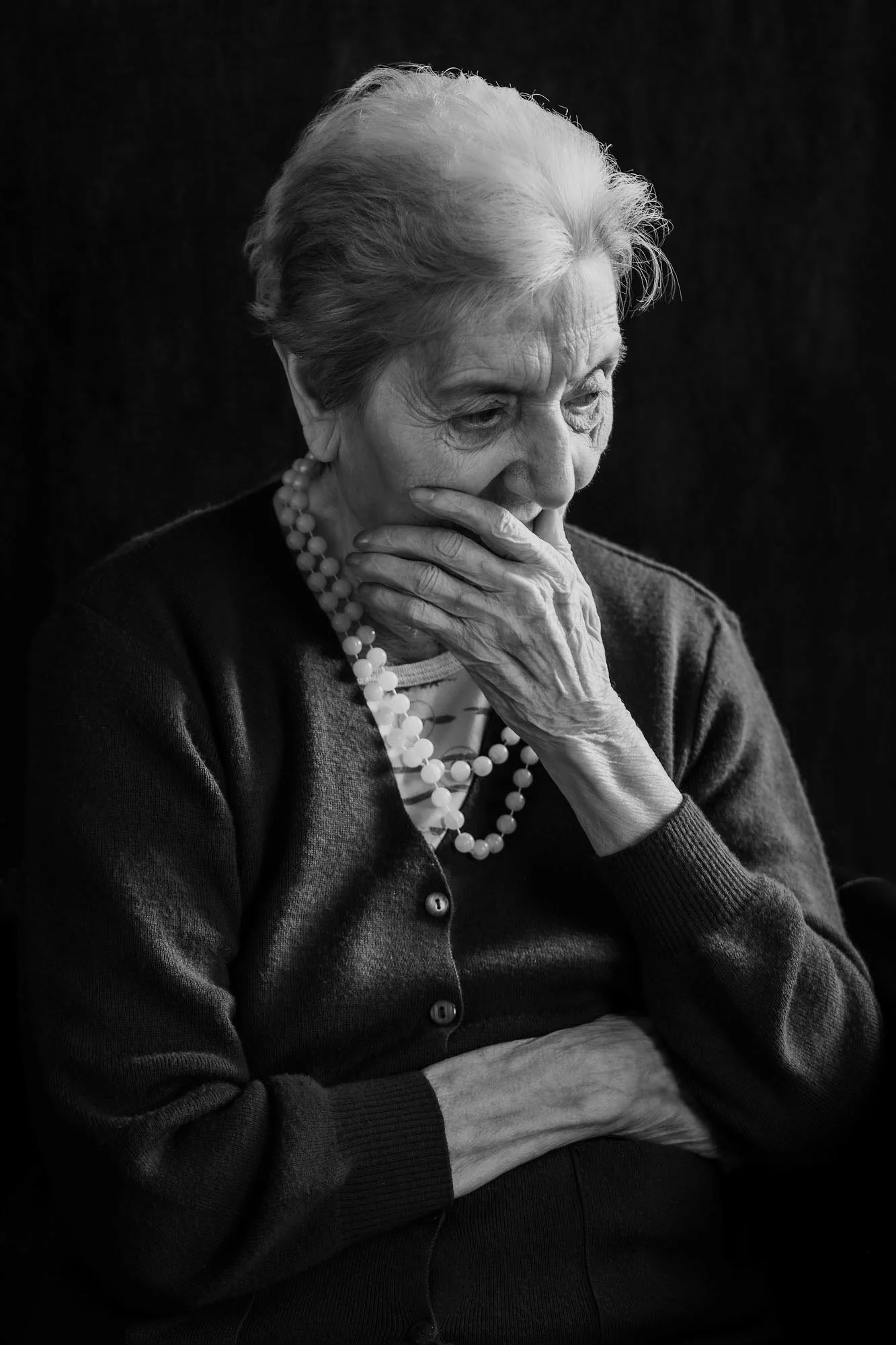 Ritratto di una donna anziana con capelli bianchi, che appare pensierosa con una mano sul viso, indossa un cardigan e una collana di perle.