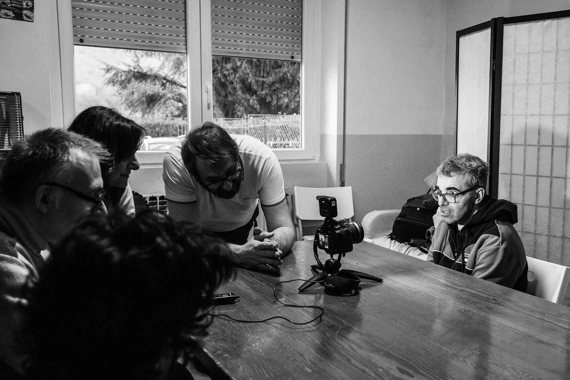 Un gruppo di persone sedute attorno a un tavolo, alcune di spalle e altre rivolte verso la telecamera, alcune stanno parlando o ascoltando, e c'è una macchina fotografica posizionata al centro del tavolo. L'immagine è in bianco e nero e mostra una sc