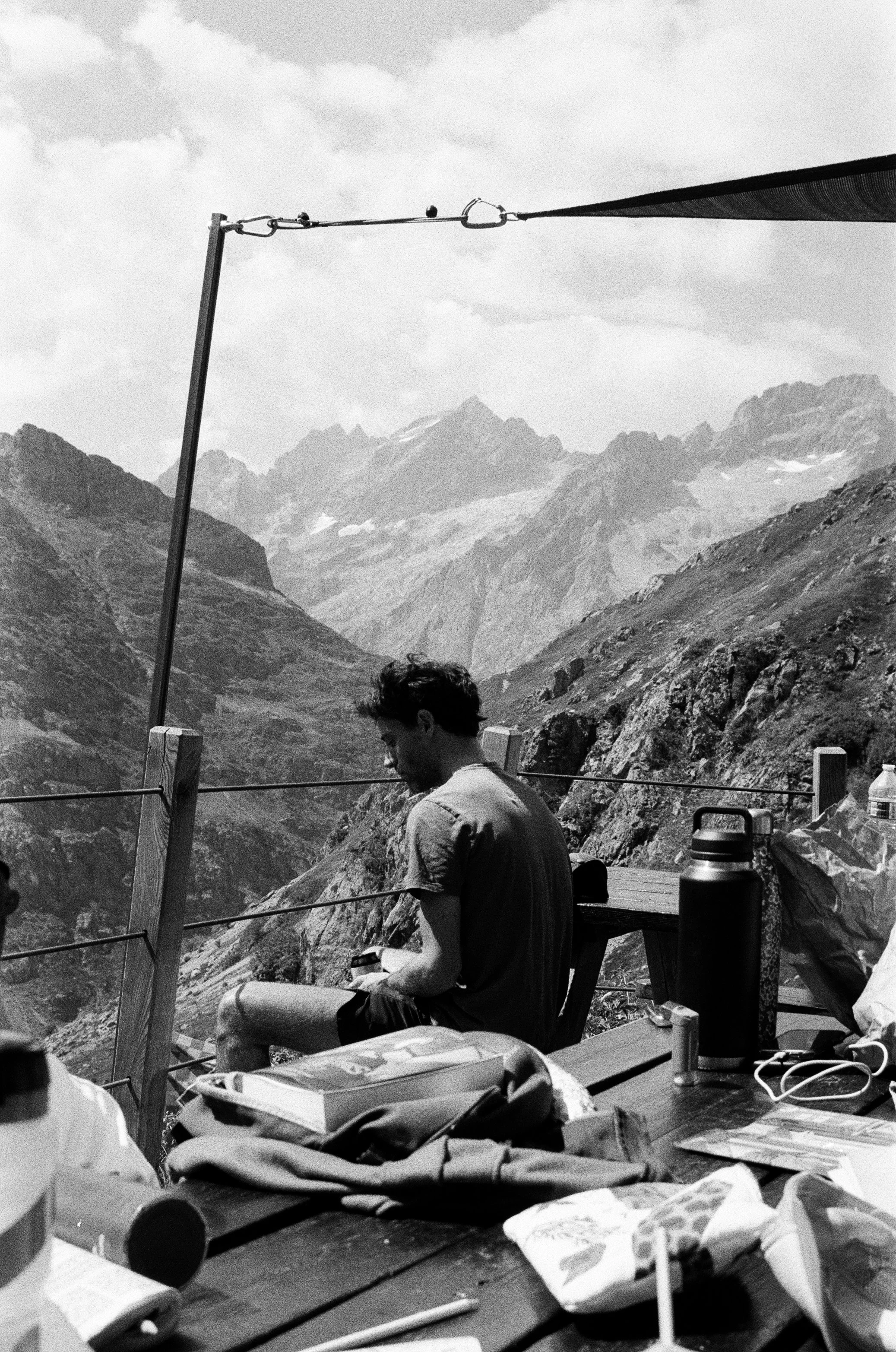 Un jeune homme assis sur une banquette en bois en altitude, entouré de montagnes, avec un sac et une bouteille thermos sur la table devant lui.
