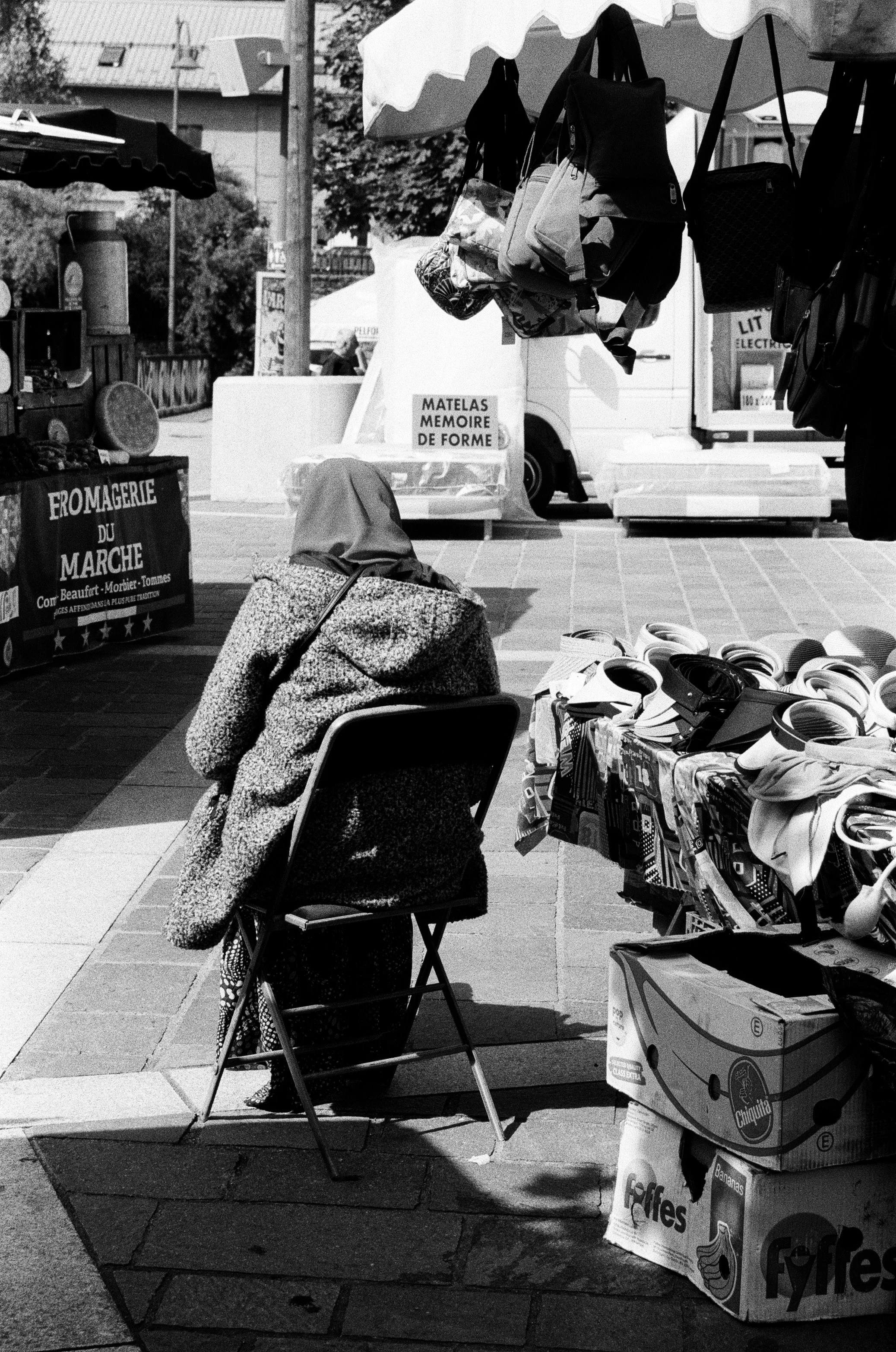 Une femme assise sur une chaise à un marché en plein air, entourée de stands vendant des sacs, des lunettes de soleil, du fromage et d'autres produits. Il y a des panneaux avec des inscriptions en français, indiquant la vente de matelas et de fromage