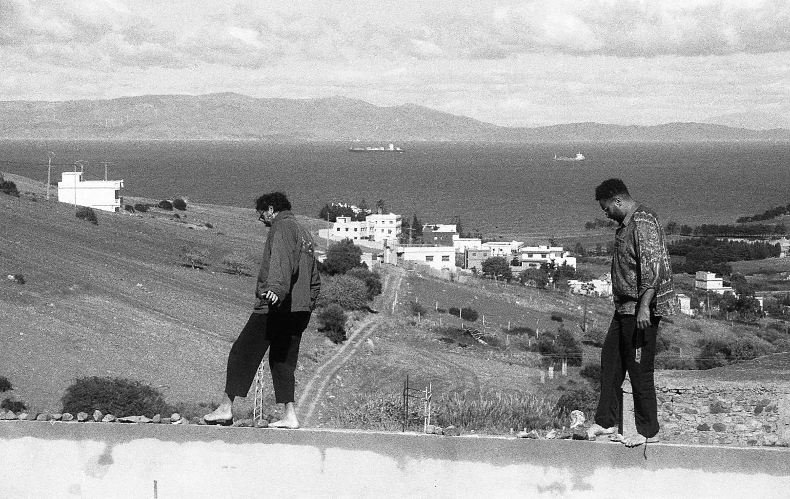 Deux personnes marchant sur une route en zone rurale avec des maisons en arrière-plan et la mer au loin, en noir et blanc.