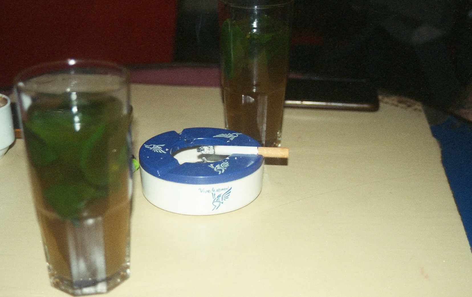 Deux verres avec des boissons vertes à la menthe, un cendrier blanc avec un mégot, et un verre avec une boisson brune, sur une table en bois.