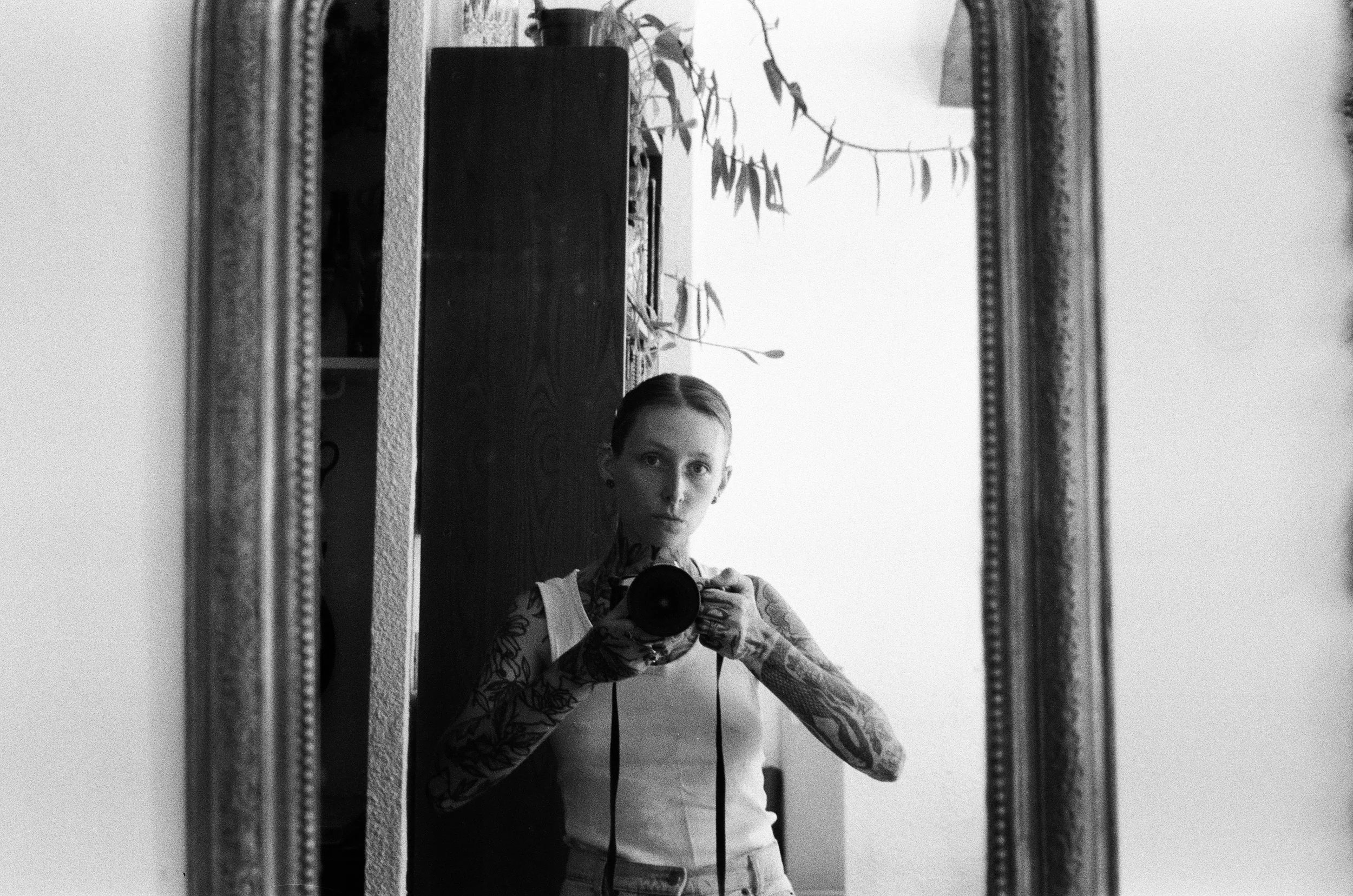 Un selfie dans un miroir montrant une femme avec des tatouages, avec un décor simple comprenant un tableau mural et quelques plantes.