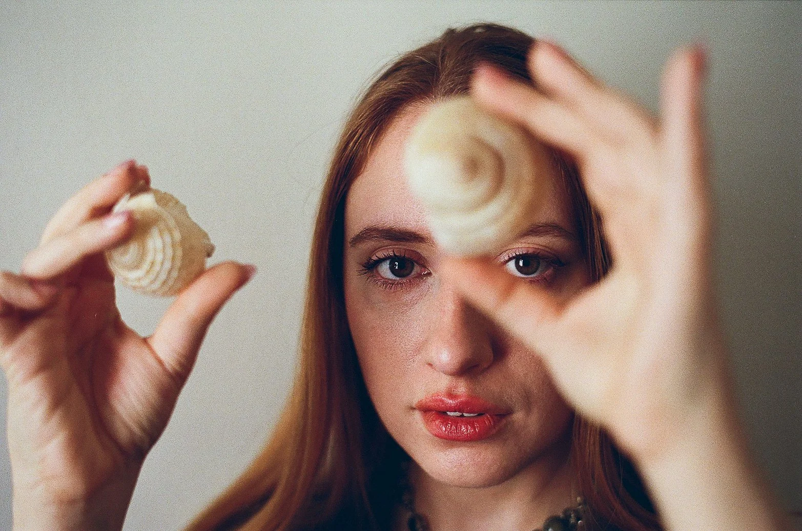 Jeune femme aux cheveux roux, tenant une coquille de mer et faisant un cercle avec ses doigts, regardant intensément l'objectif.