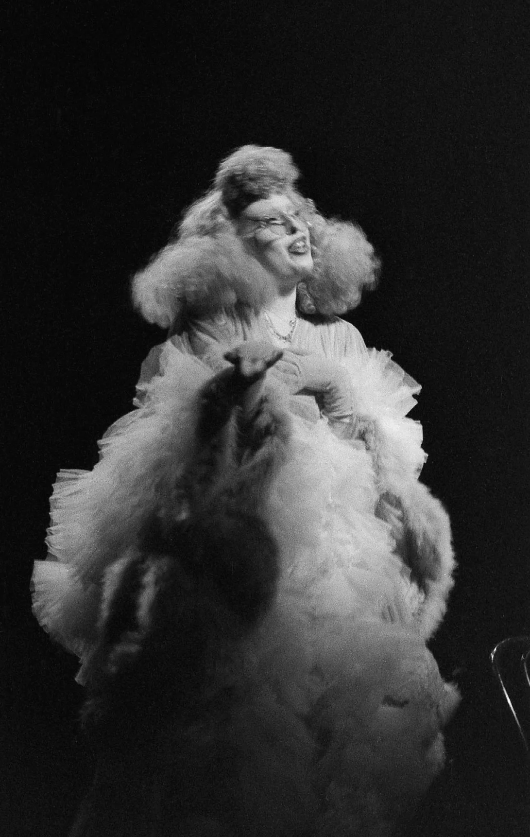 Femme blonde portant une robe volumineuse et un chapeau pelucheux, souriant, dans un décor sombre.