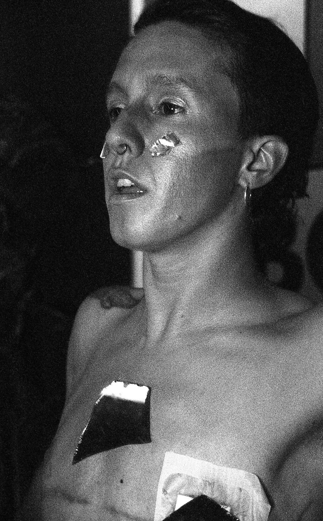 Une femme avec des tatouages, un piercing au nez, portant un earring, regardant vers la gauche, en noir et blanc.
