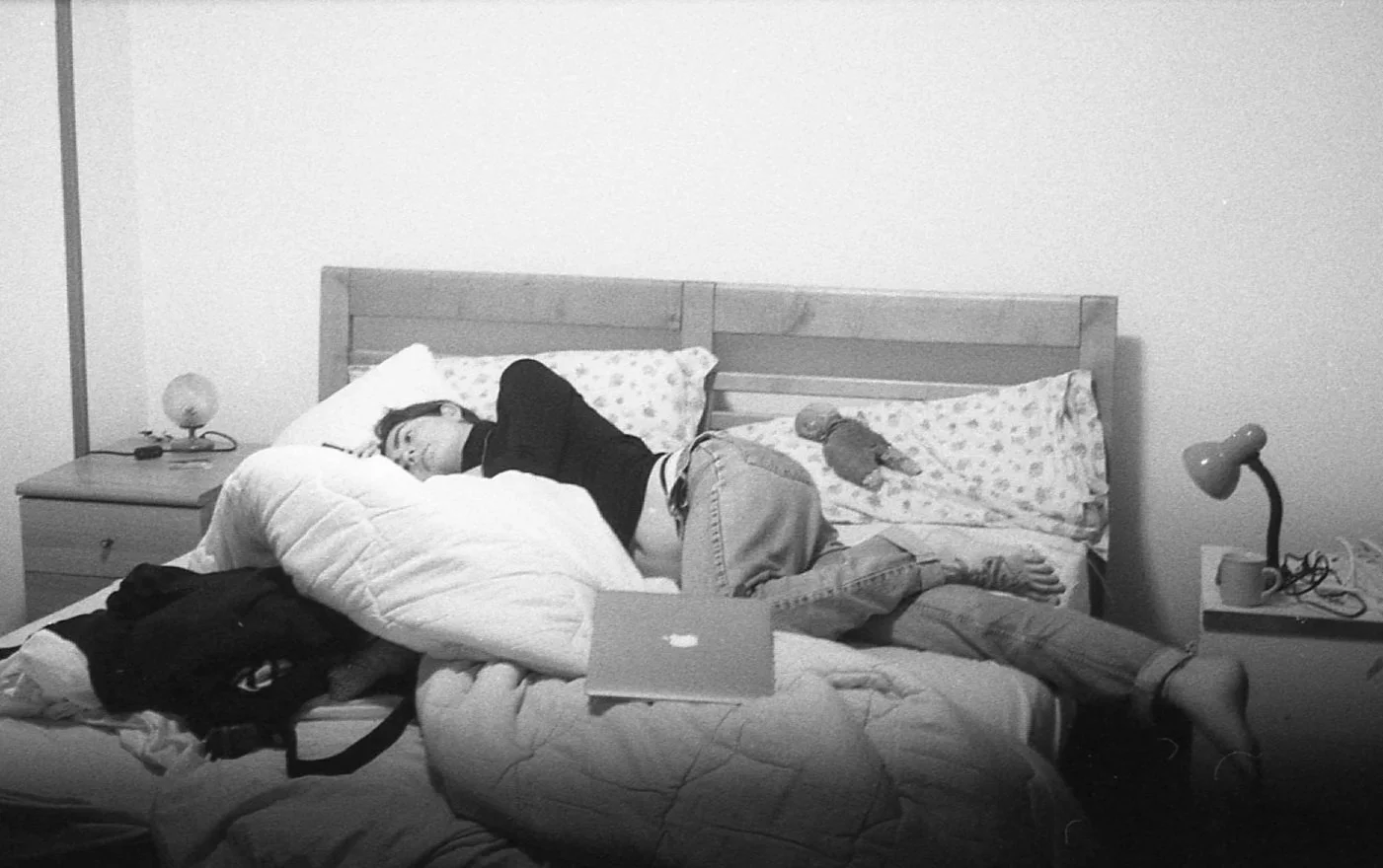 Une personne dort sur un lit, allongée sur le côté, dans une chambre avec deux lampes de chevet. La personne porte des lunettes, un sweat noir, un pantalon clair, et a un kit de tennis. Un doudou en peluche est posé sur l'oreiller, le lit n'a pas de 