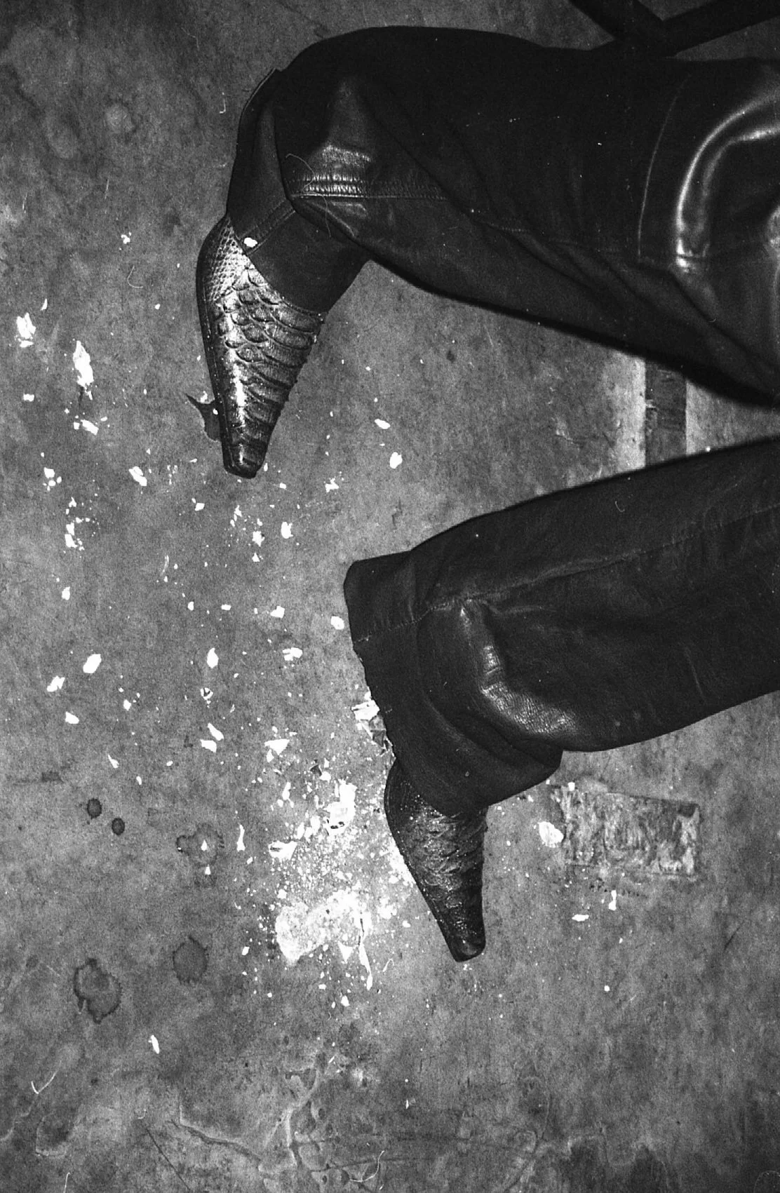Chaussures pointues en cuir, portant des pantalons en cuir, vu de dessus sur un sol sale avec des débris et une tache de peinture.