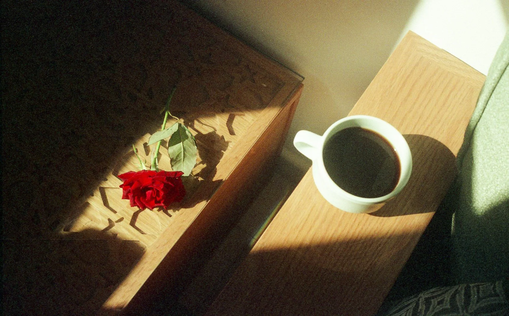 Une tasse de café noir sur une table en bois à côté d'une rose rouge allongée avec des feuilles vertes, sous un éclairage naturel.