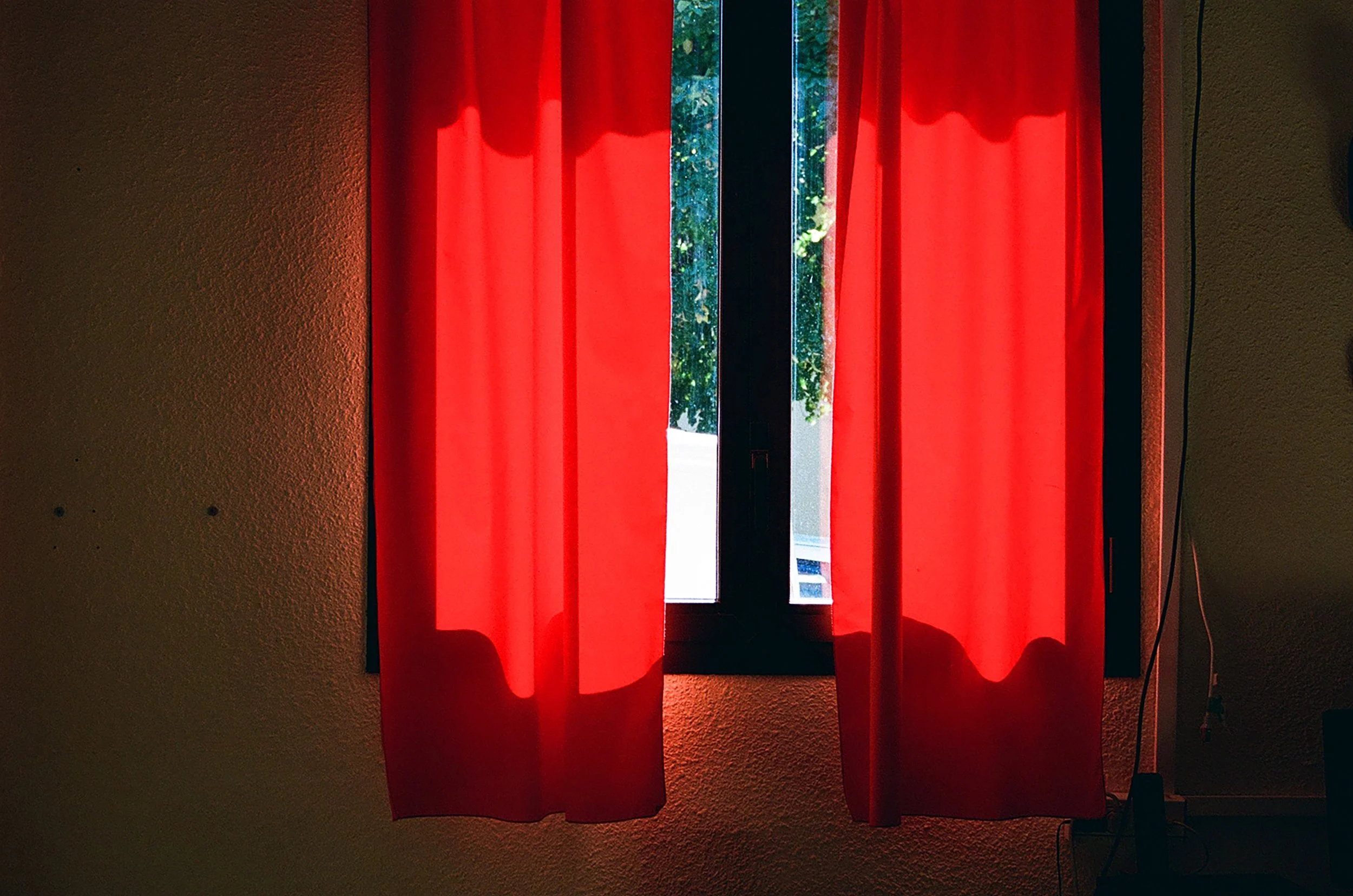 Fenêtre avec rideaux rouges laissant passer la lumière, vue sur l'extérieur avec des arbres verts.