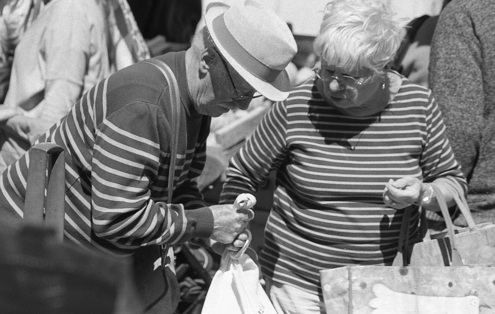 Deux personnes âgées discutant et échangeant des objets dans un marché, portant des vêtements à rayures, l'une avec un chapeau. Image en noir et blanc.