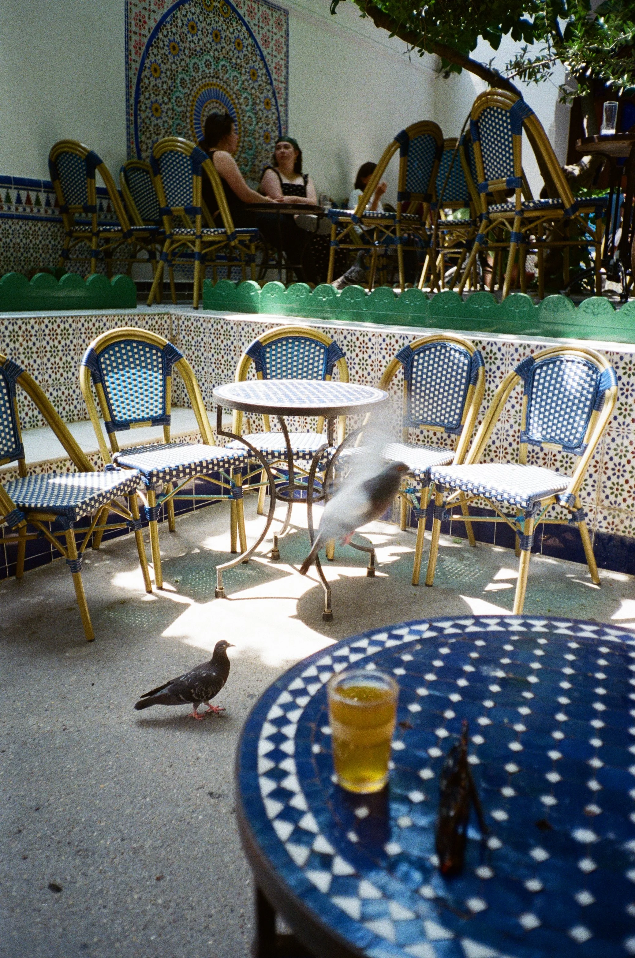 Une terrasse de café avec des chaises en rotin et des tables à motif de mosaïque bleue et blanche. Deux pigeons se trouvent au sol, et un verre avec une boisson jaune est posé sur une table. En arrière-plan, des personnes assises discutent, avec un m