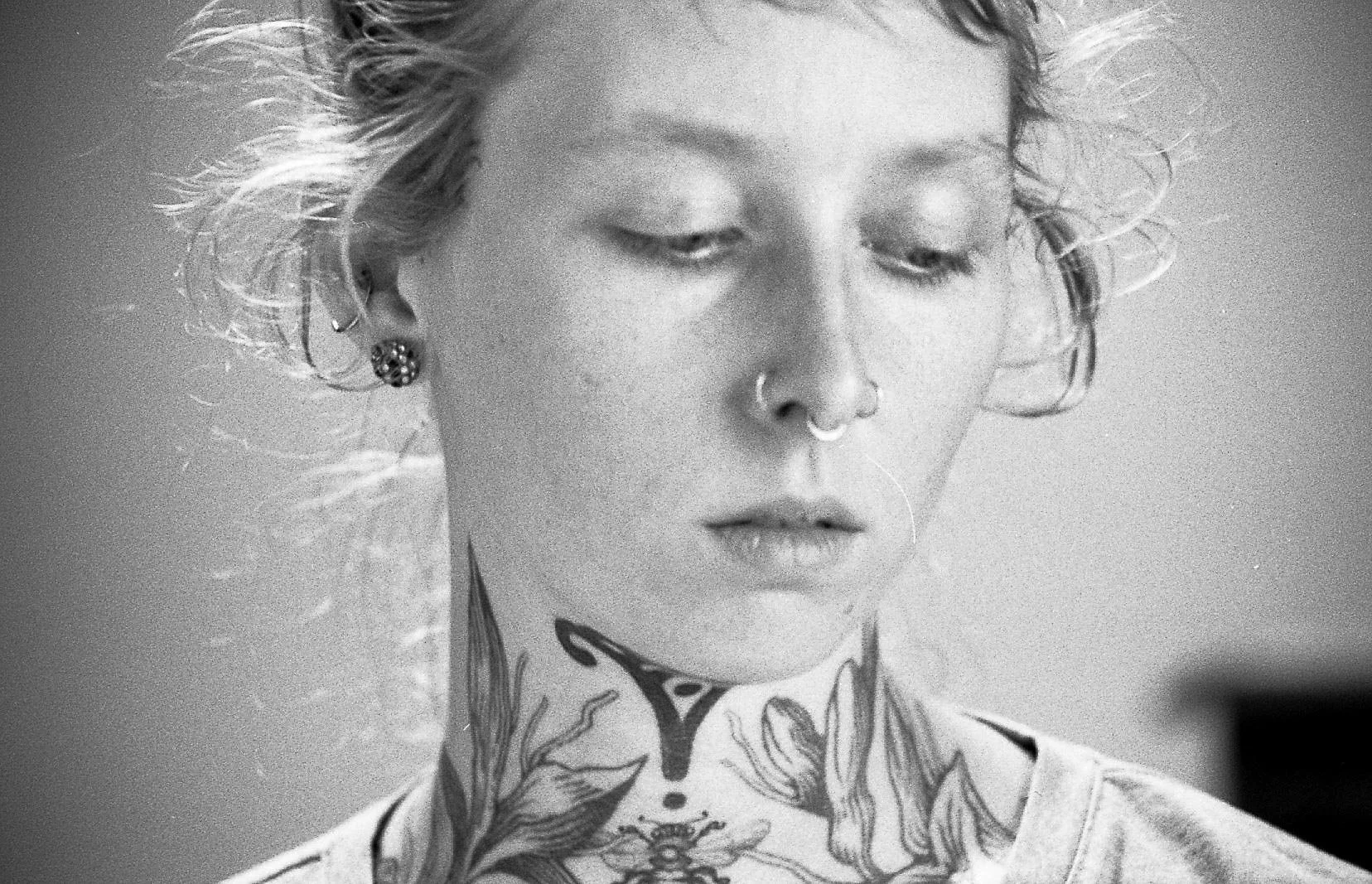 Portrait en noir et blanc d'une jeune femme avec des cheveux en bataille, plusieurs piercings, tatouages visibles sur le cou et la poitrine, regardant vers le bas.