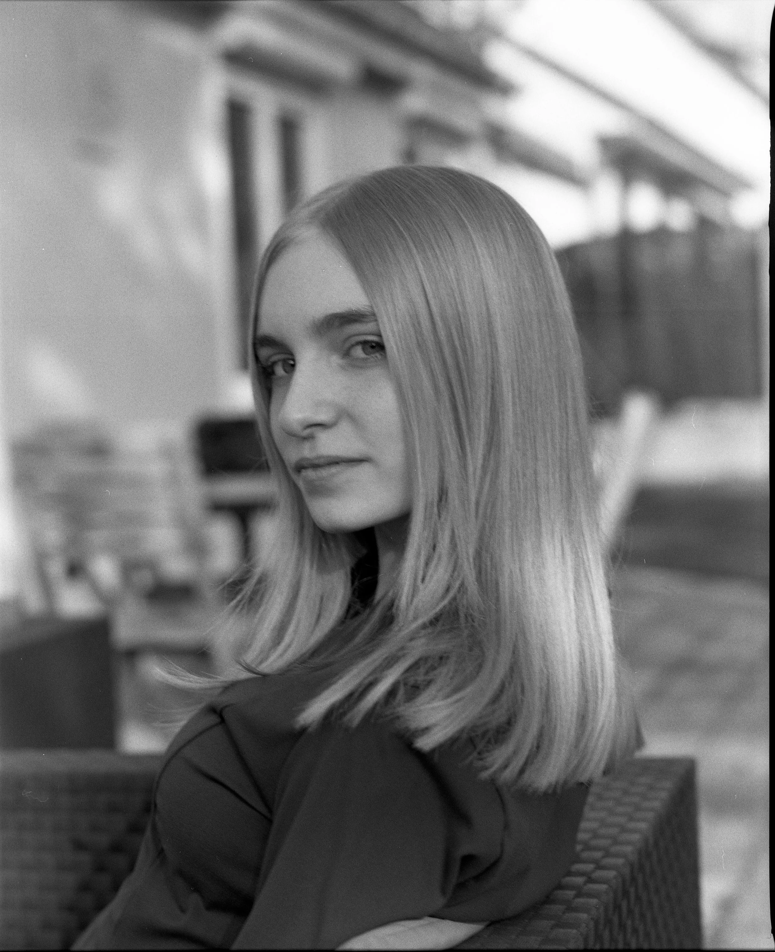 Portrait en noir et blanc d'une jeune femme aux cheveux longs et lisses, assise sur un fauteuil à l'extérieur avec un bâtiment en arrière-plan.