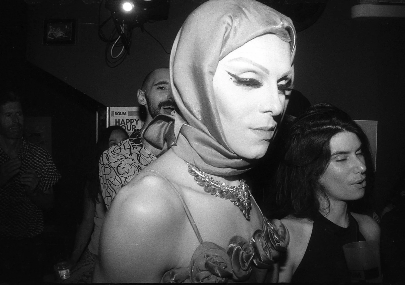 Une femme portant un foulard en satin et un collier brillant, entourée de plusieurs autres personnes dans un environnement festif.