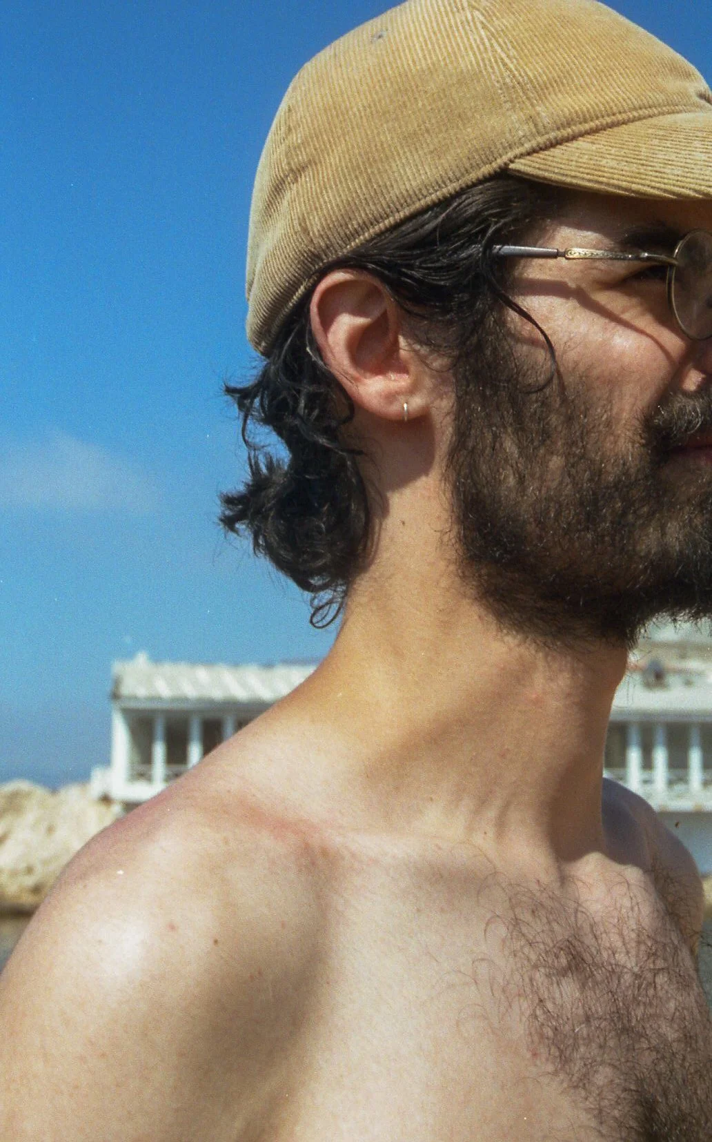Un homme avec une barbe portant une casquette beige et des lunettes, en extérieur, avec un bâtiment blanc en arrière-plan.