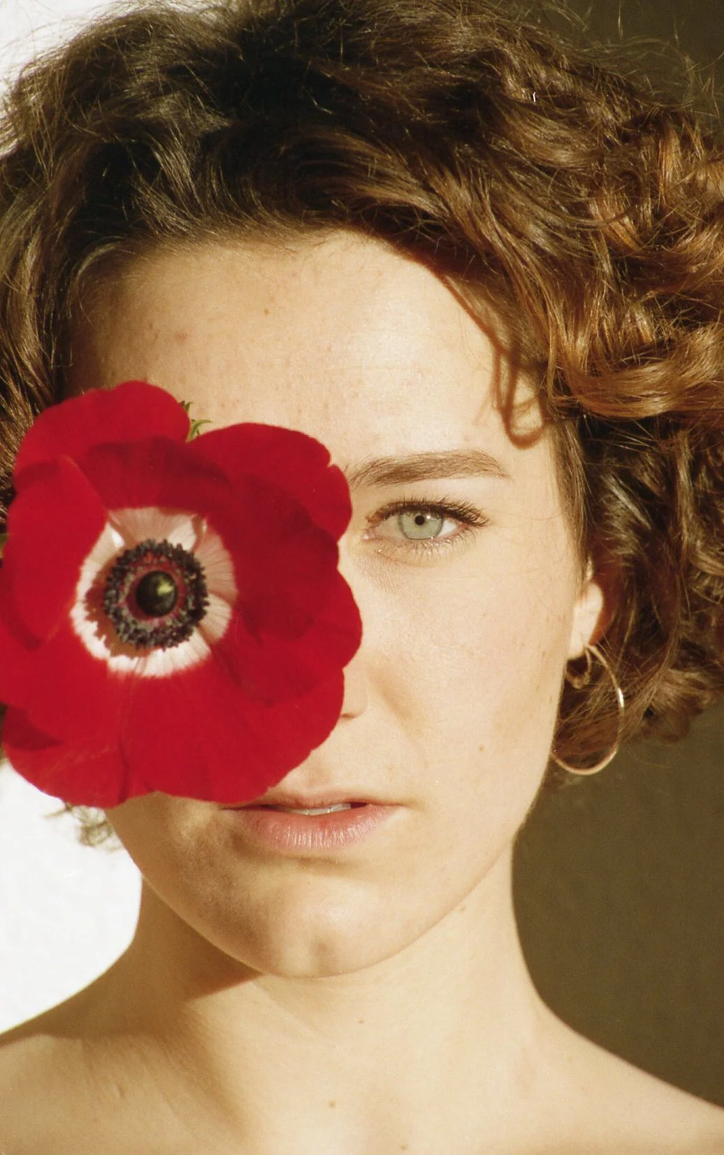 Une femme aux cheveux bouclés avec un regard vert, tenant une grande fleur rouge devant un œil, découvrant une partie de son visage.