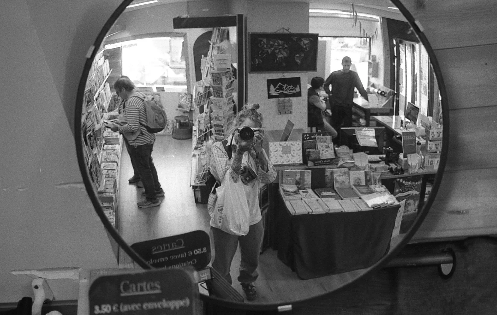 Une scène à l'intérieur d'une librairie vue à travers un miroir rond. Plusieurs personnes parcourent les rayons : une femme au centre se prend en photo avec un appareil photo, une autre à gauche lit un livre, et un homme à l'arrière se tient près d'u