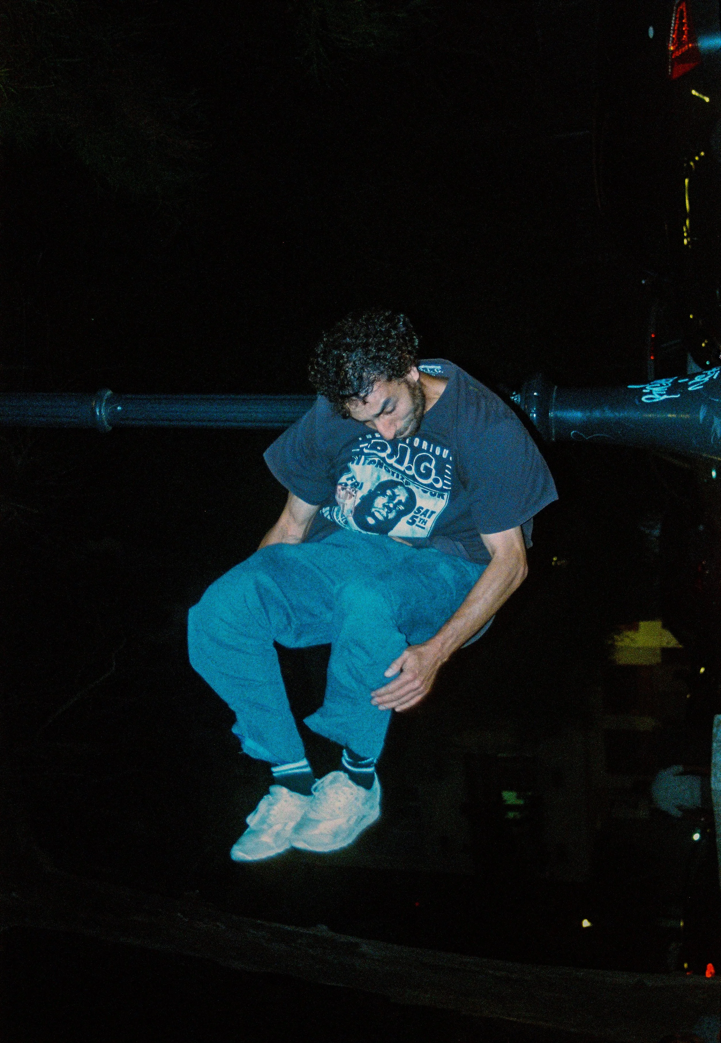 Jeune homme avec cheveux bouclés, assis sur un abri-bus, regardant vers le bas, portant un t-shirt noir avec une illustration, pantalon ample bleu, et chaussures blanches, dans un environnement nocturne.