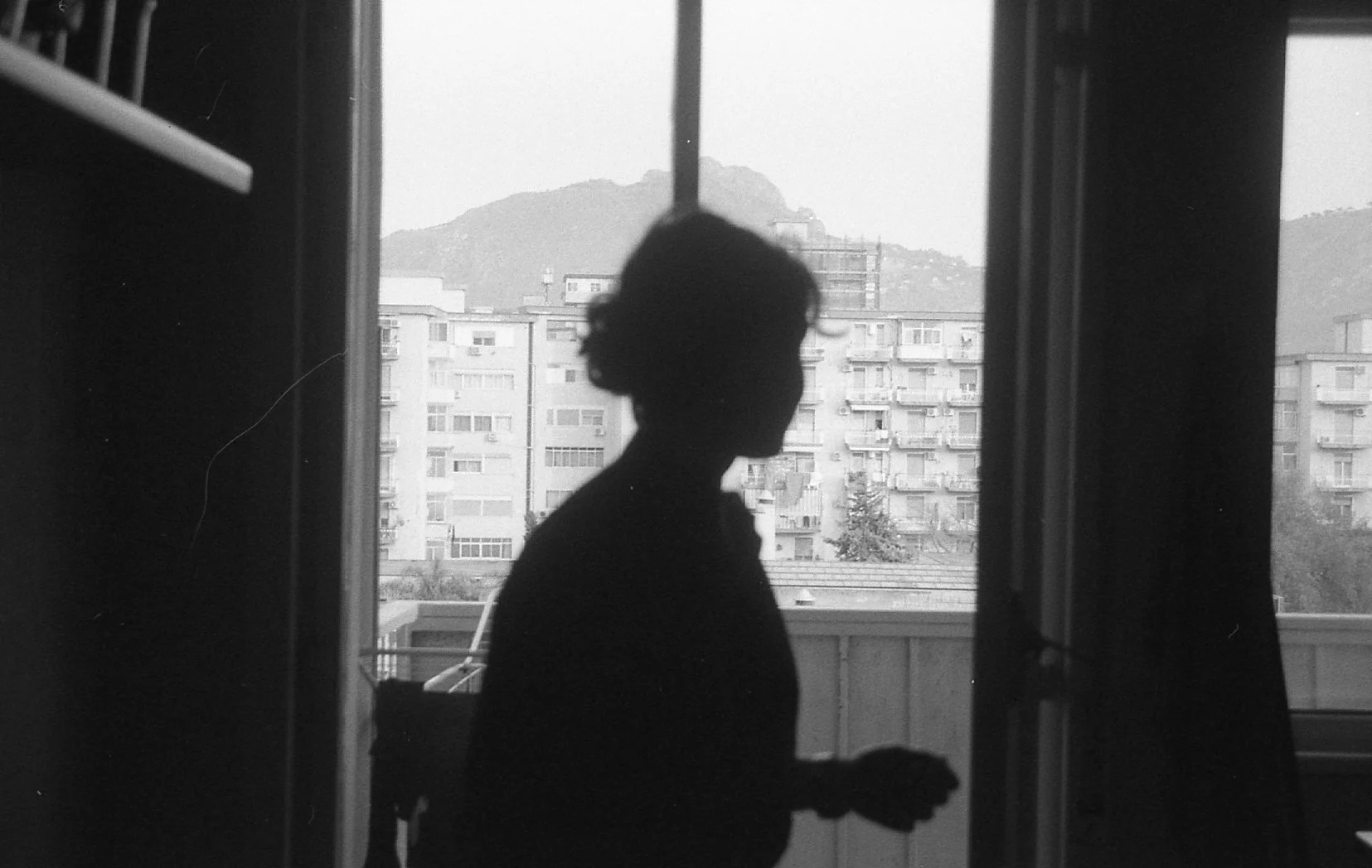 Silhouette d'une personne debout sur un balcon, vue depuis l'intérieur d'une pièce avec une fenêtre ouverte, en arrière-plan un immeuble résidentiel et des collines.