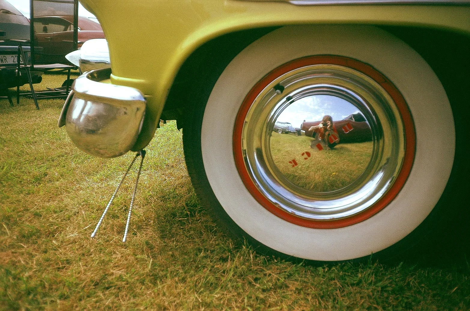 Reflet dans un pneu de voiture vintage jaune avec une jante blanche, montrant une femme prenant une photo, entourée d'un champ avec des voitures en arrière-plan.