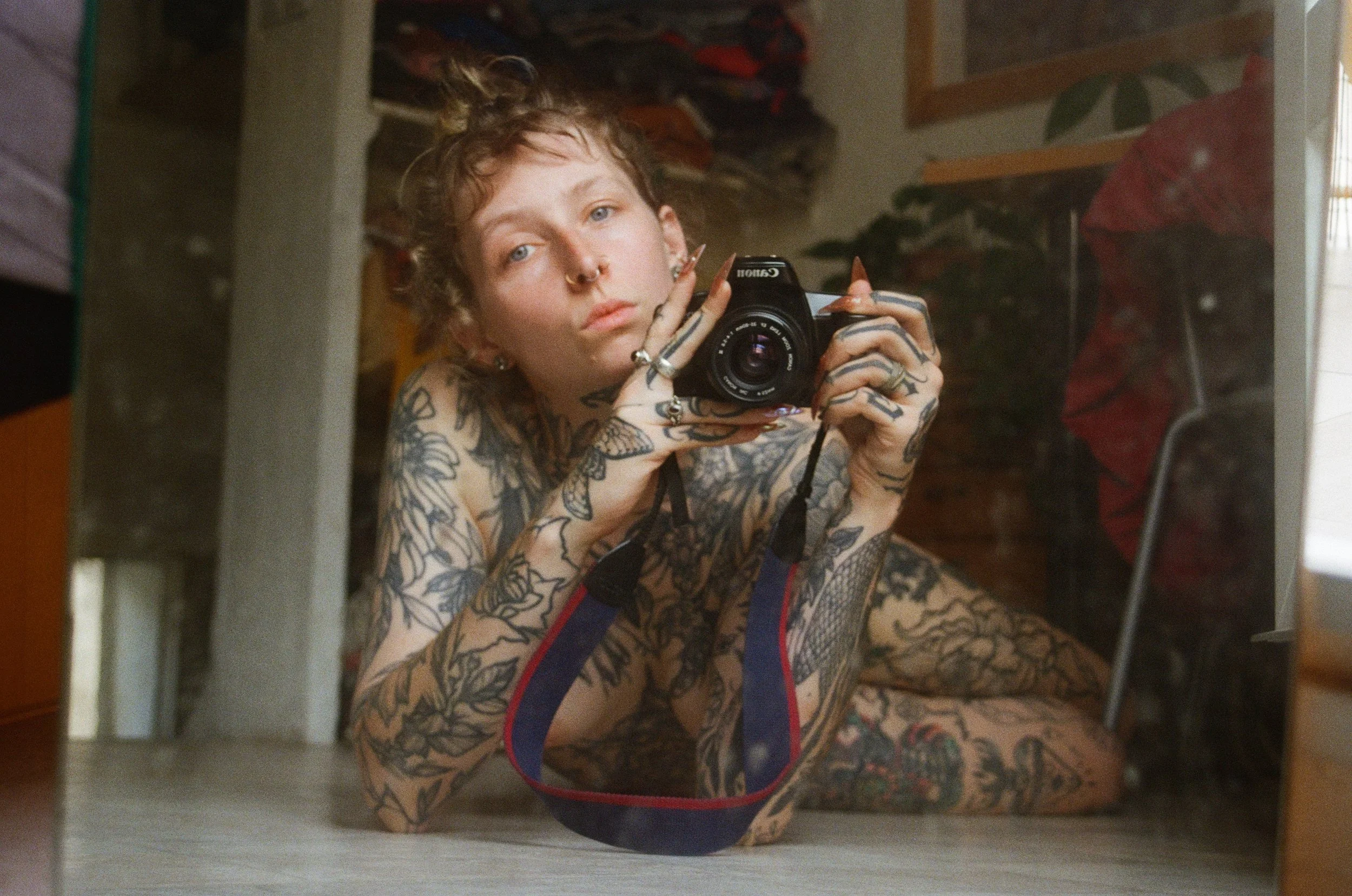 Jeune femme avec tatouages, cheveux en désordre, prenant une photo dans un miroir, portant un slip en résille et un sweat-shirt, dans une pièce avec des étagères et des objets en arrière-plan.